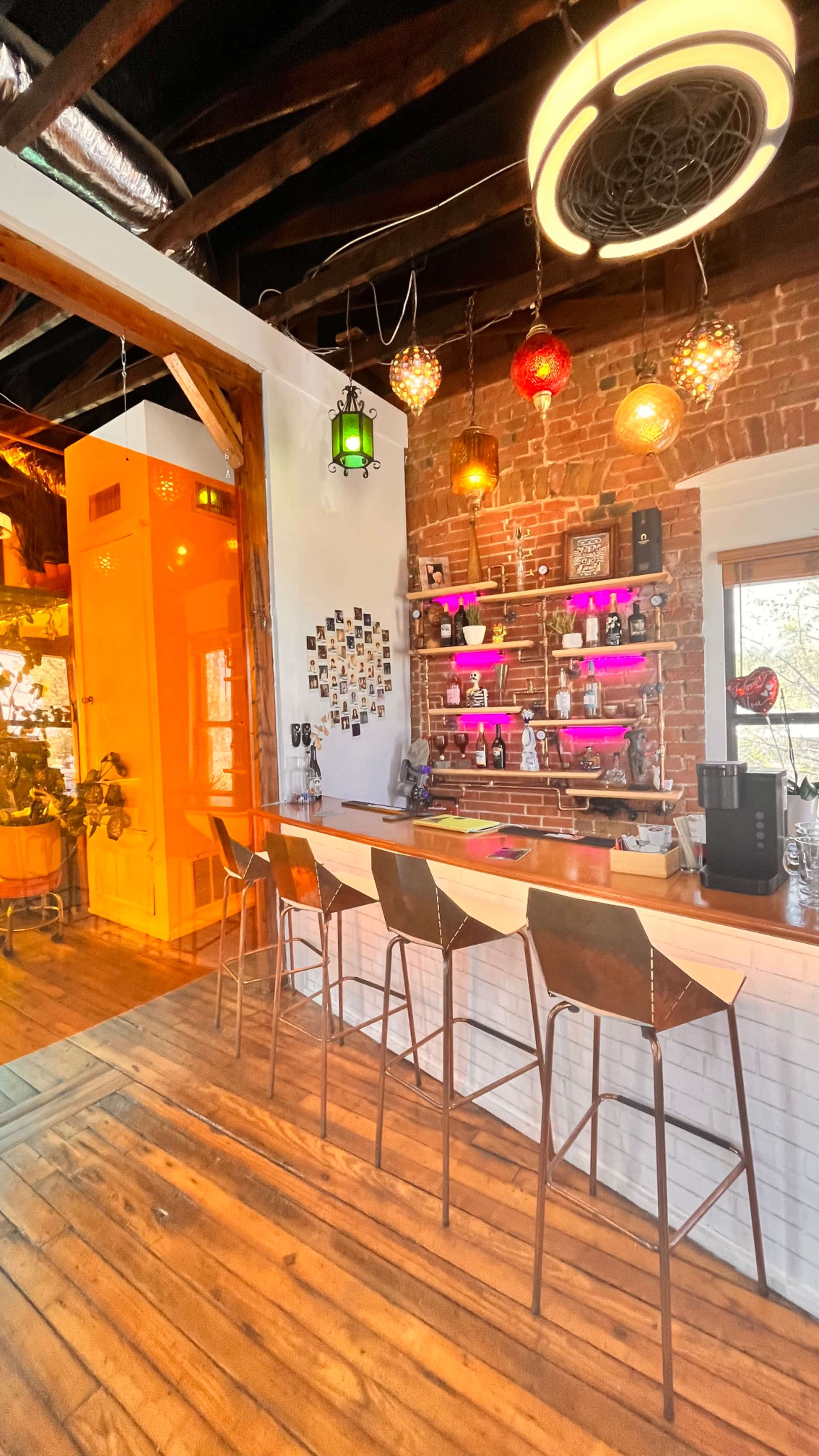 A modern bar area featuring a long wooden countertop with high stools, set against a wall of exposed brick adorned with colorful lights and decorative items.