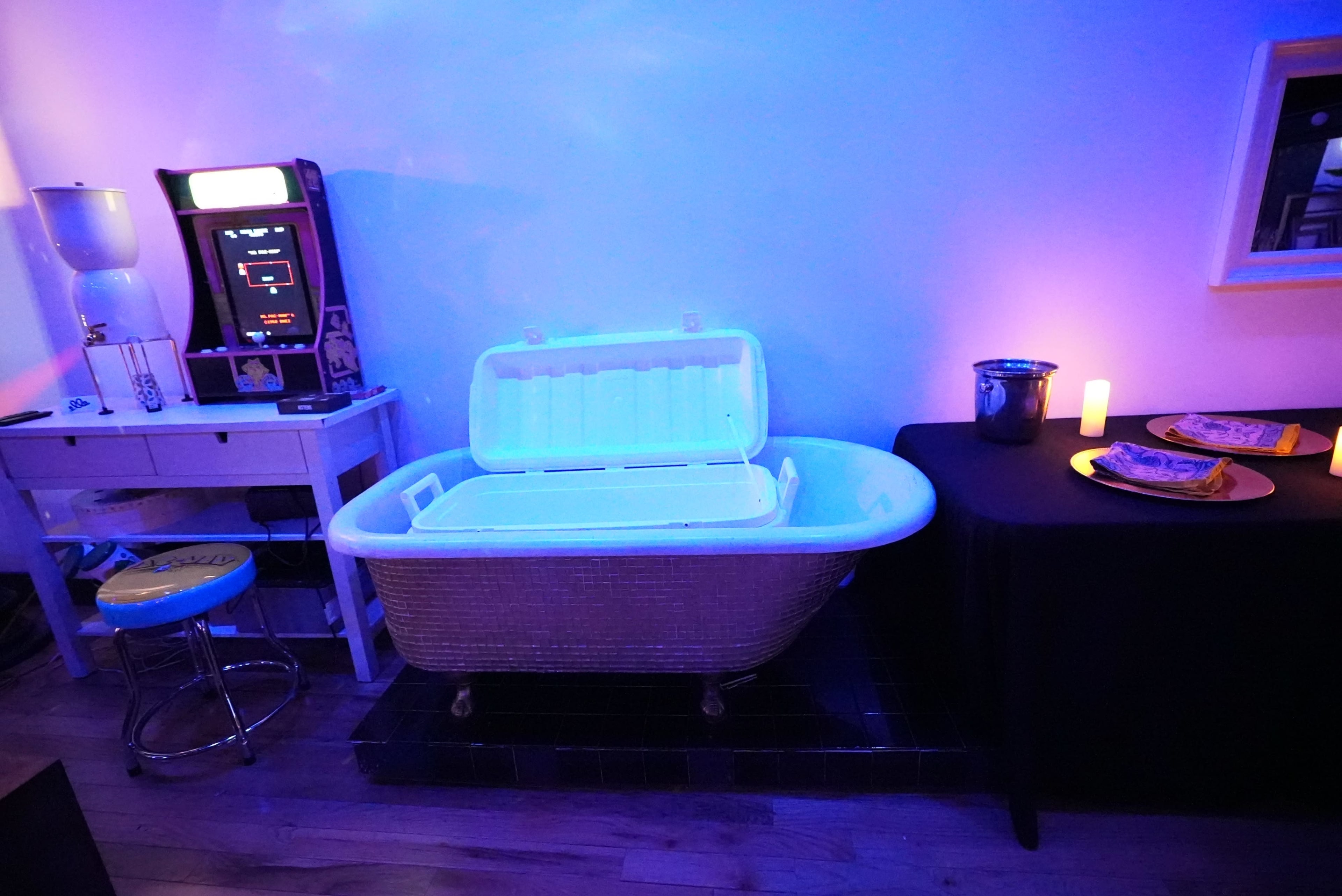 The image shows a white vintage bathtub with its lid open, positioned next to a candlelit table with pizza, a drinks bucket, and a retro arcade machine in a dimly lit room.