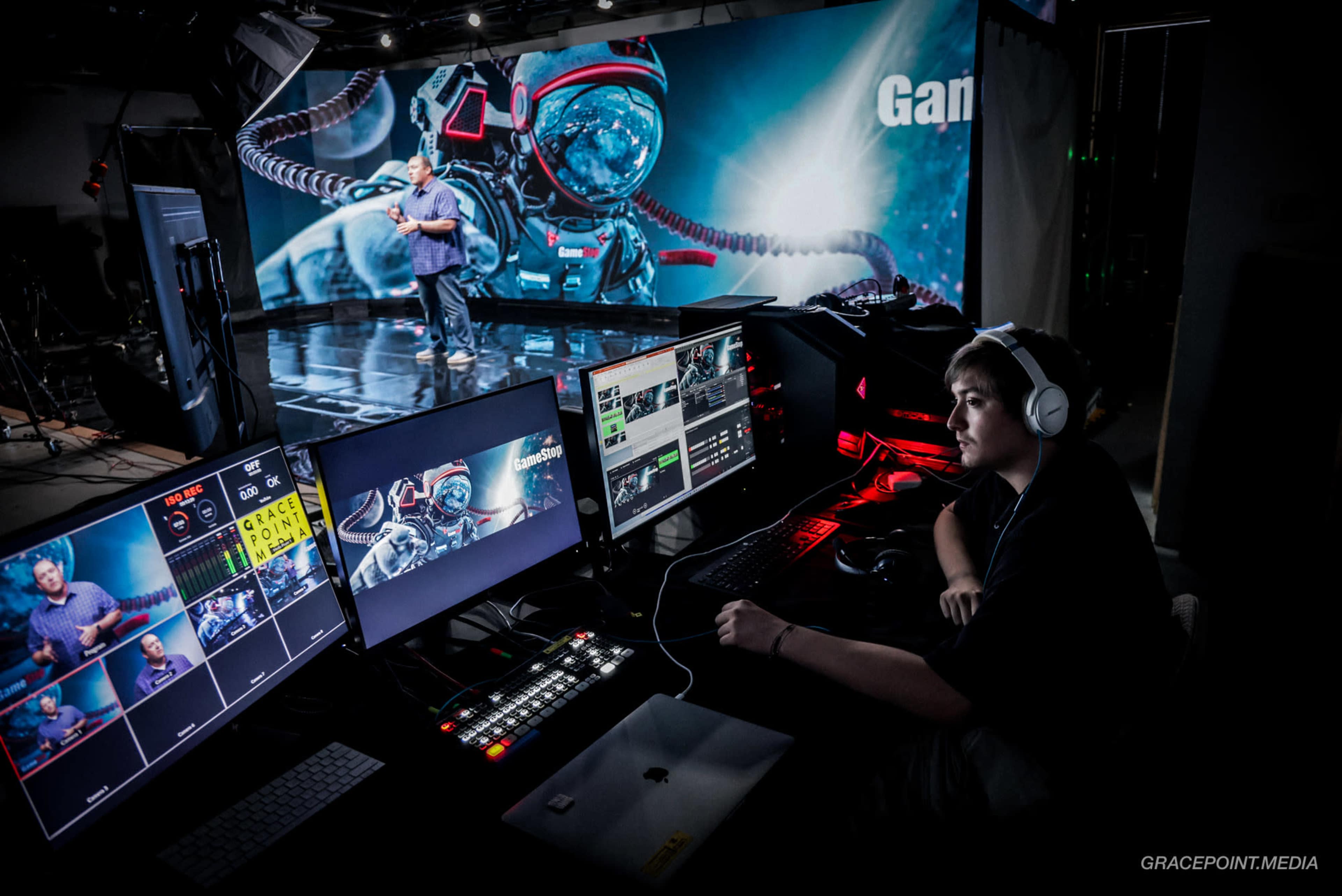A person operates a media control station with multiple screens while a presenter speaks on stage in front of a large digital backdrop featuring a space-themed graphic.