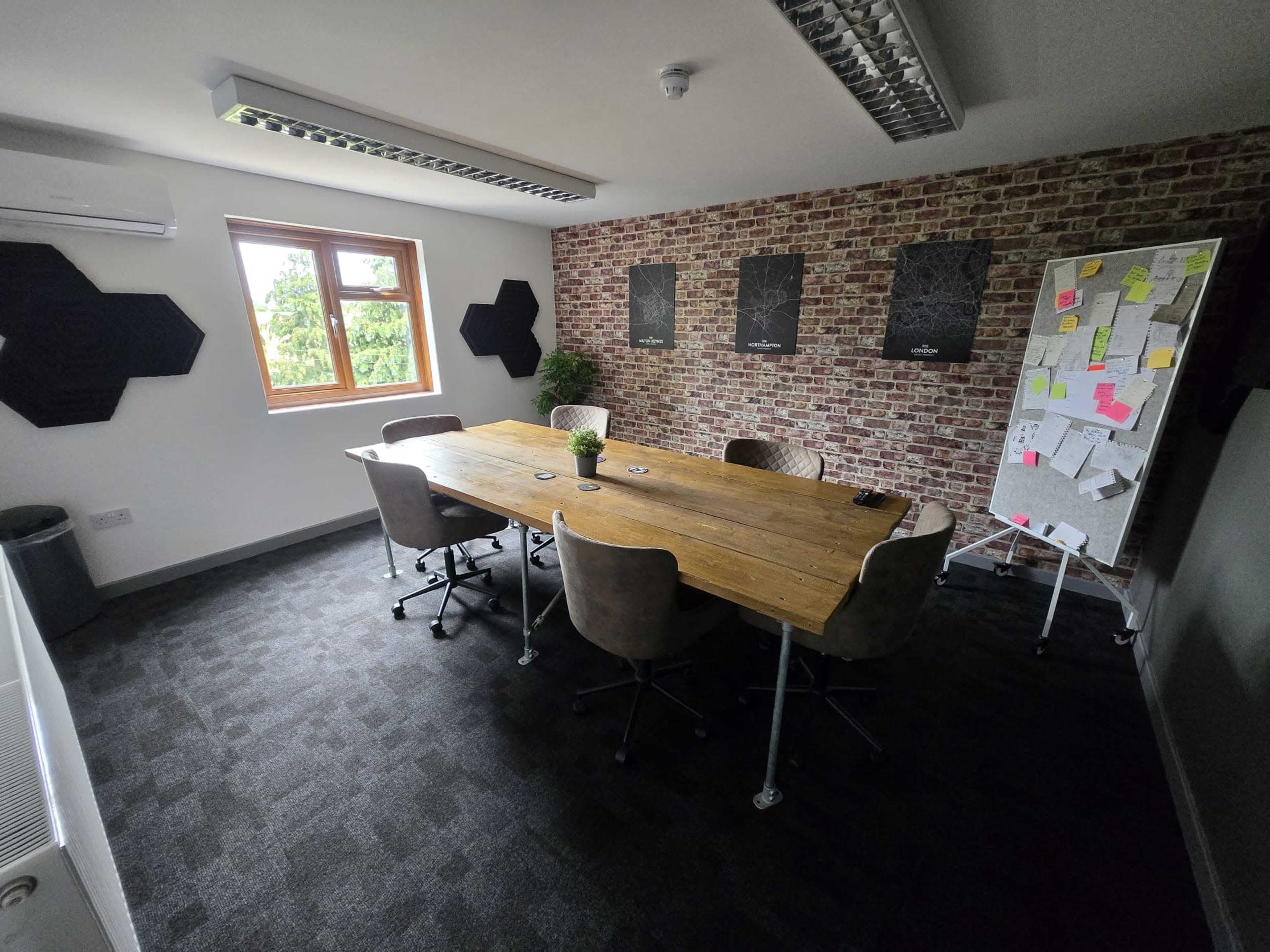 The image shows a modern meeting room with a large wooden table surrounded by six office chairs, brick walls, and a notice board covered with colorful papers.