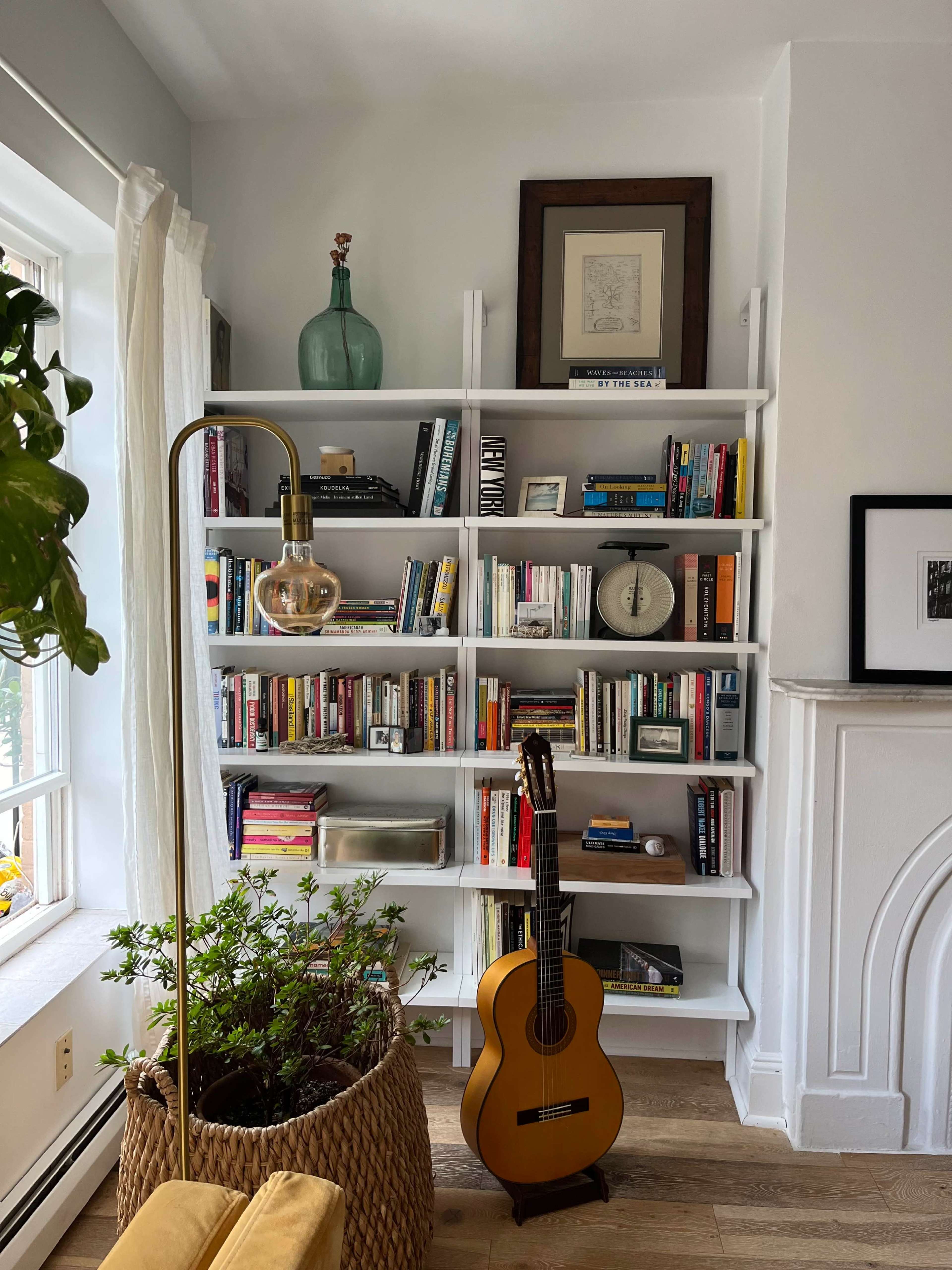 A white bookshelf filled with various books and decorative items stands next to a potted plant and a guitar in a well-lit room.