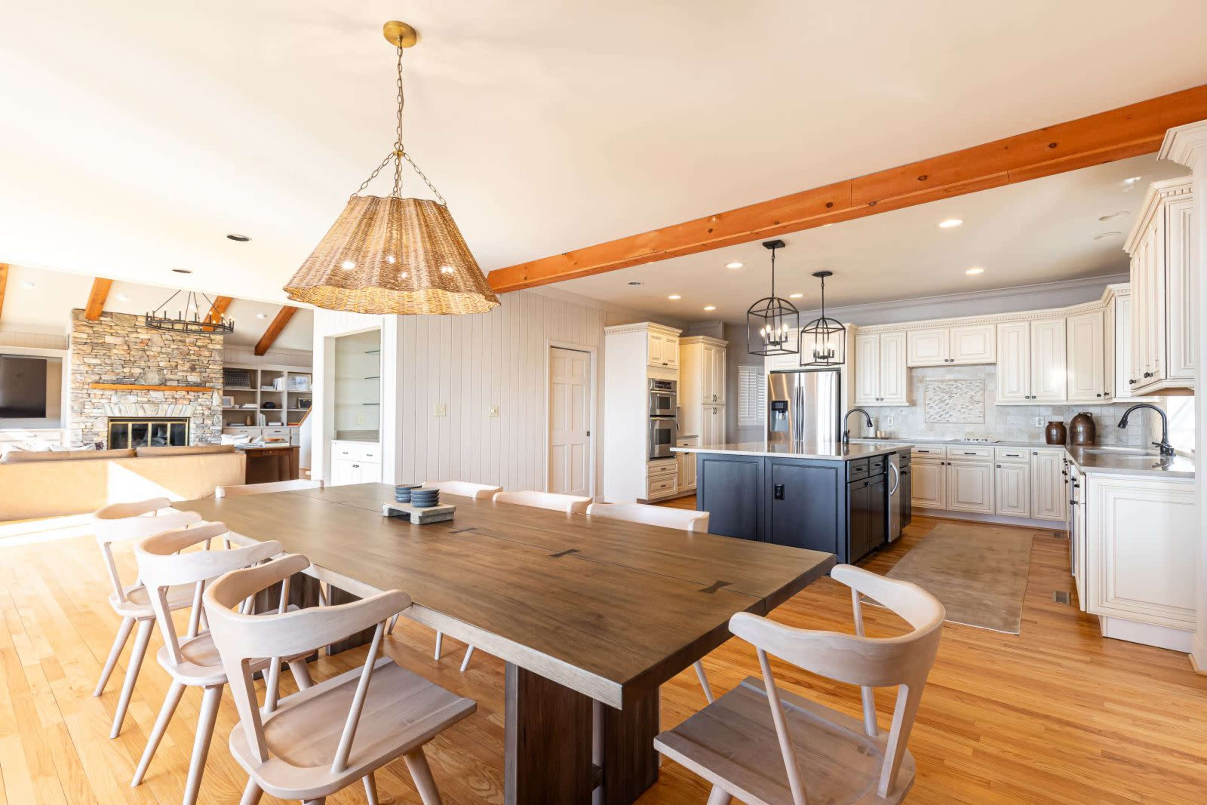 A spacious kitchen features a large wooden dining table surrounded by eight chairs, with a stone fireplace visible in the background.