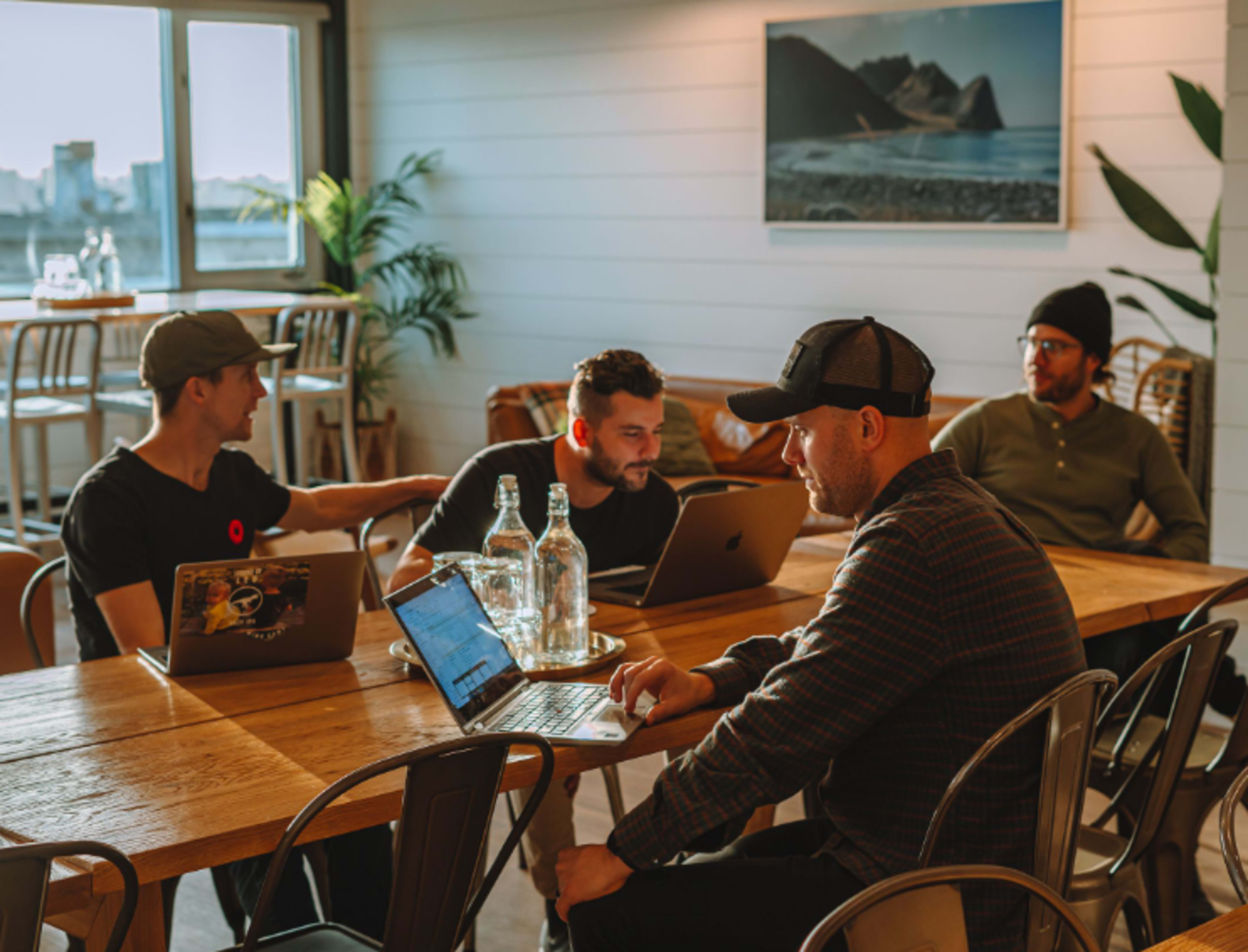 Four individuals work together at a wooden table in a bright, modern workspace with laptops and water bottles.