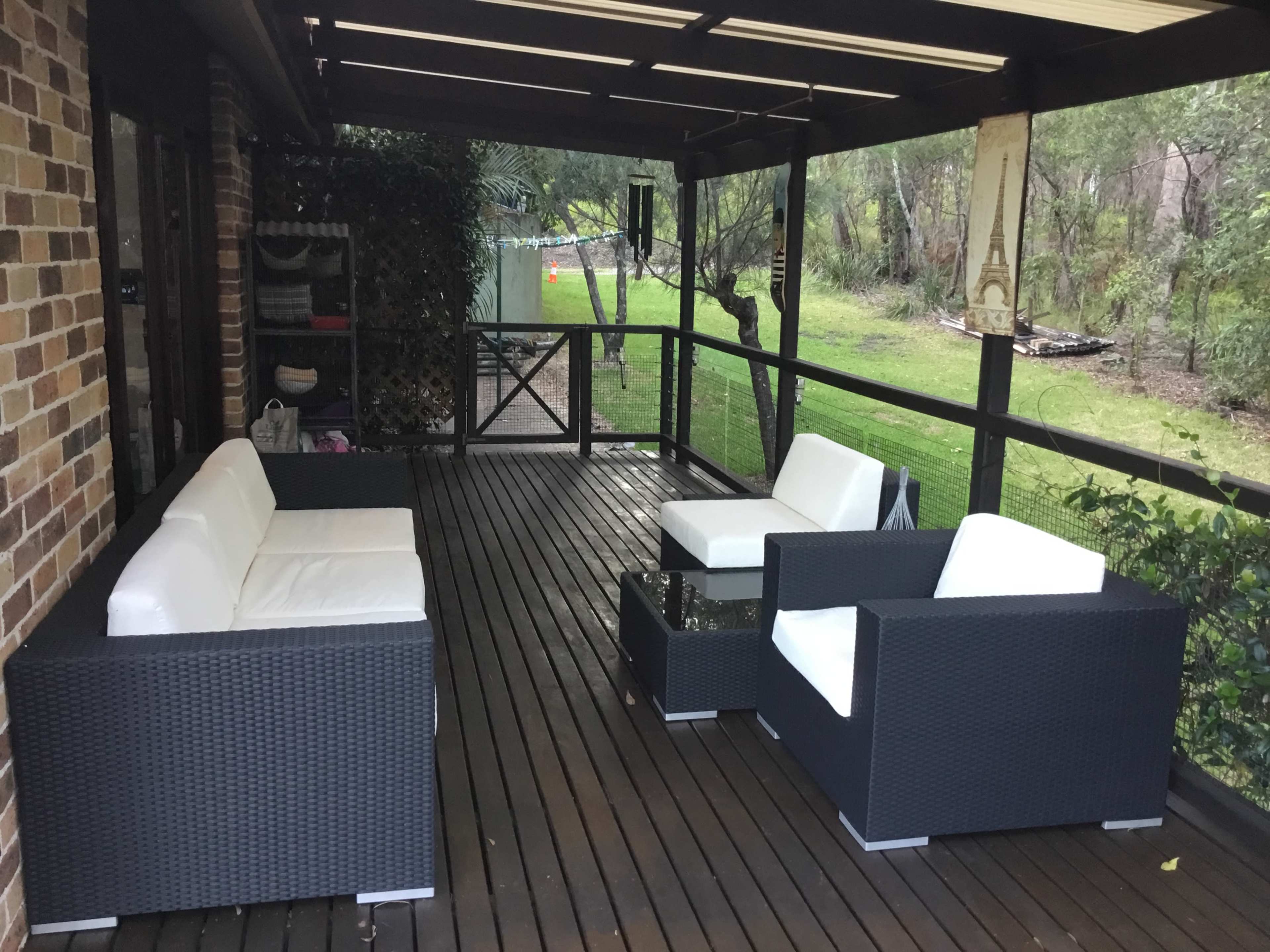 A covered outdoor patio furnished with two white sofas and a glass-top coffee table, surrounded by greenery.