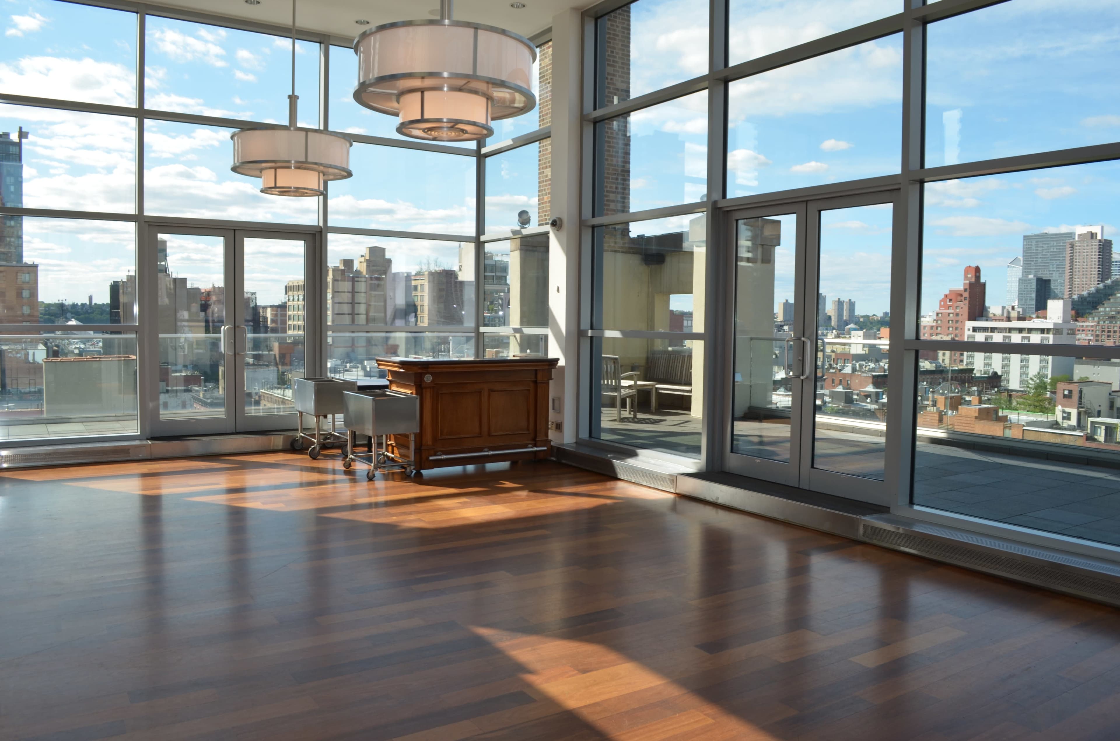 The image shows a spacious, well-lit room with large windows offering a view of a city skyline and wooden flooring.