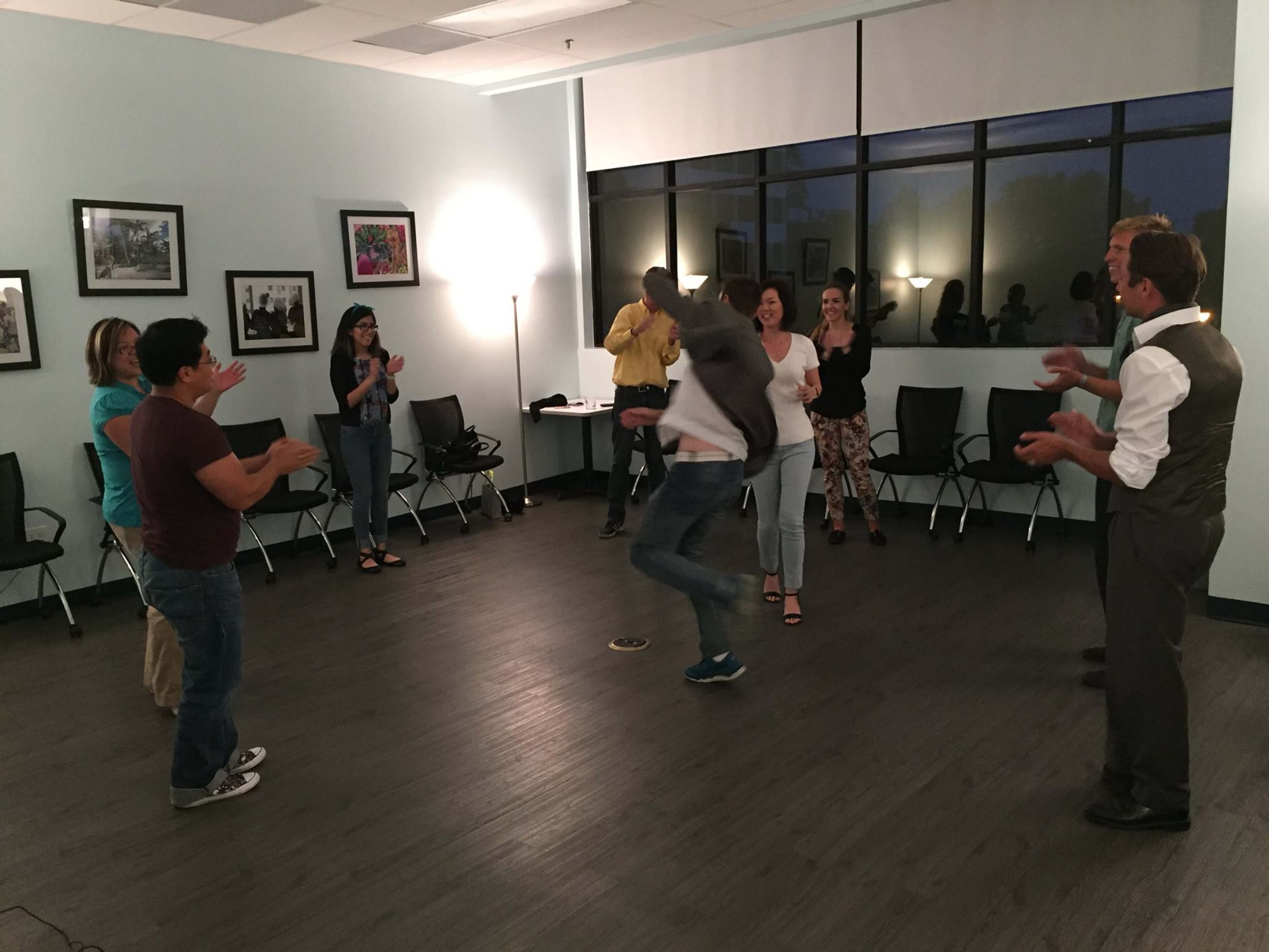 A group of people is clapping and watching as one individual jumps in the center of a well-lit room.