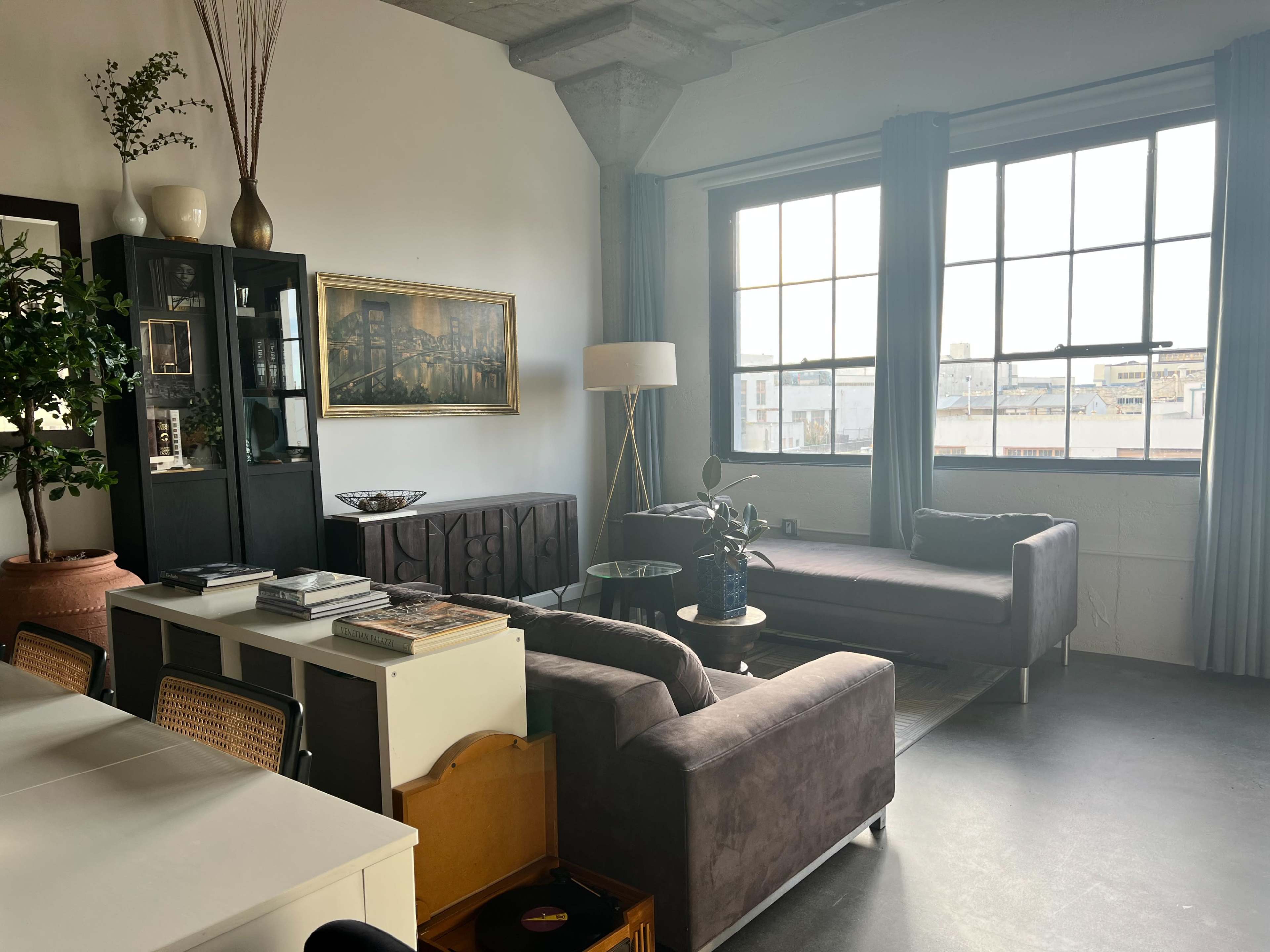The image shows a contemporary living room with large windows, a dark sofa, a light-colored armchair, a dining table, and various decorative plants.