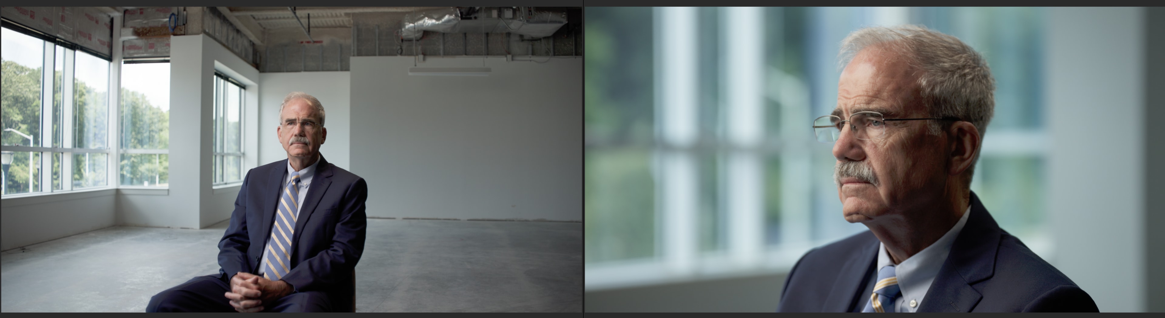A man in a suit sits on a chair in an empty room with large windows.