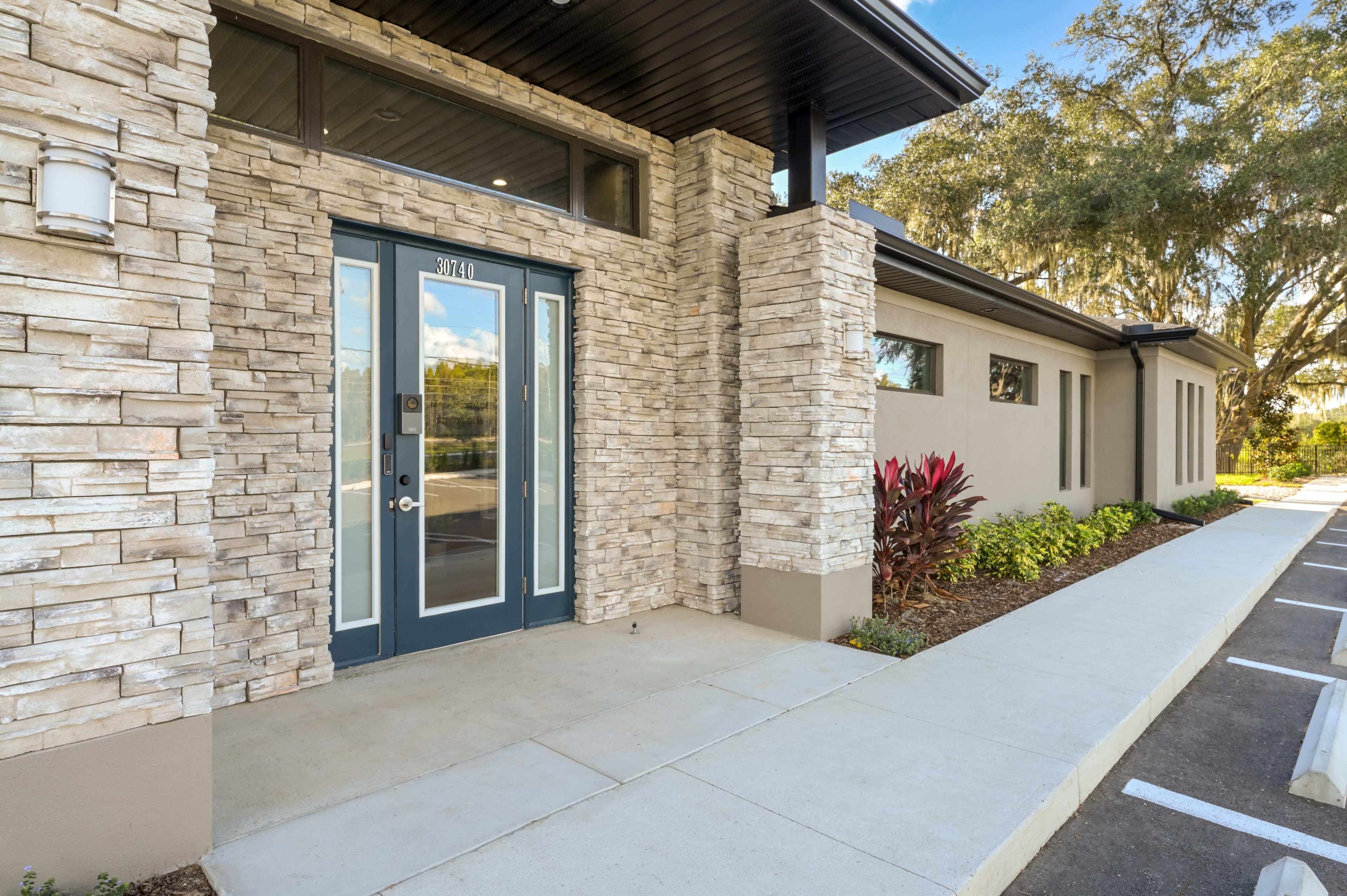 The image shows a modern building entrance with a stone facade, a blue door, and a concrete walkway beside landscaped greenery.