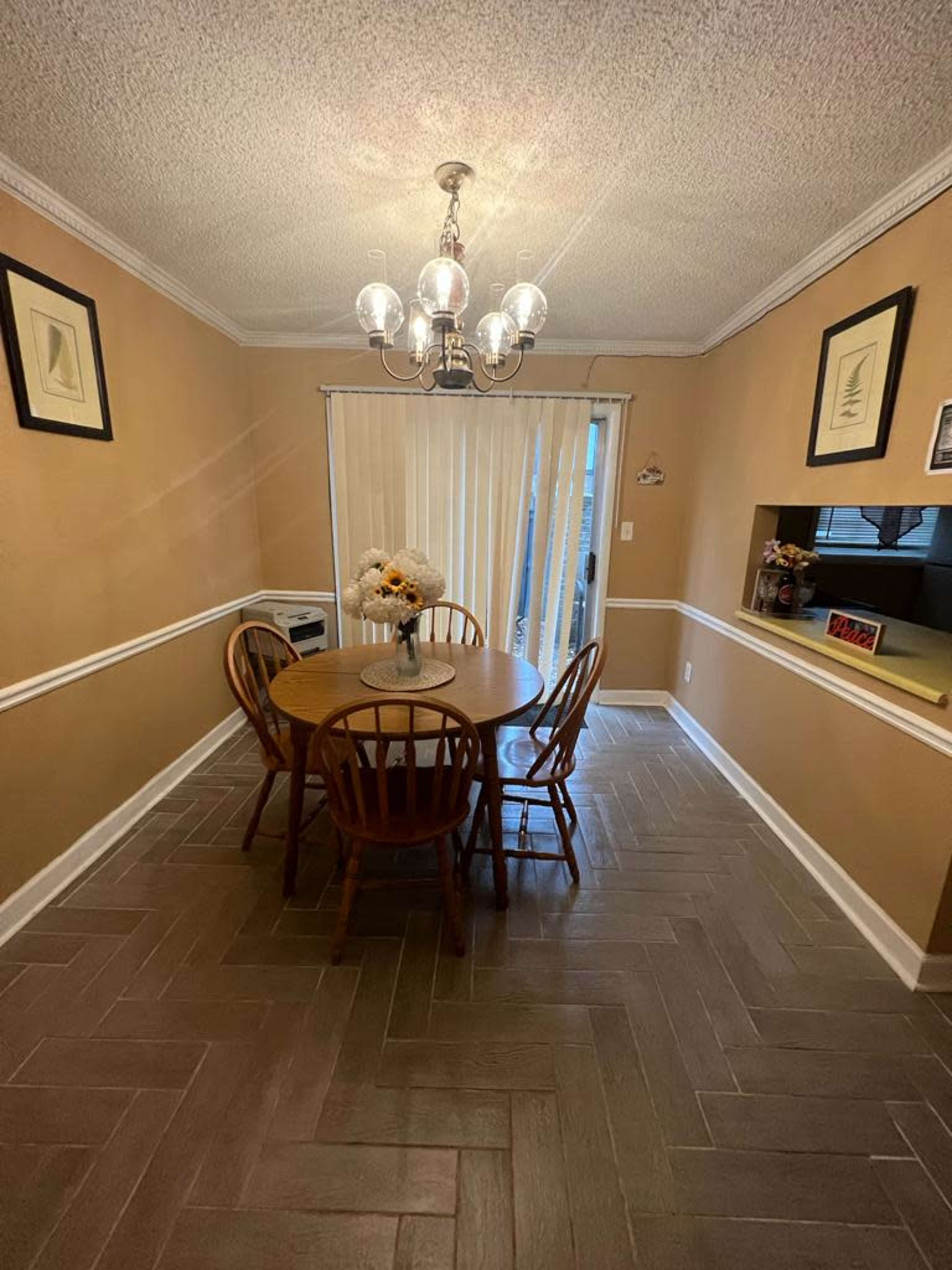 A round wooden dining table with four chairs is positioned in a softly lit room with beige walls and a chandelier overhead, leading to a sliding glass door.