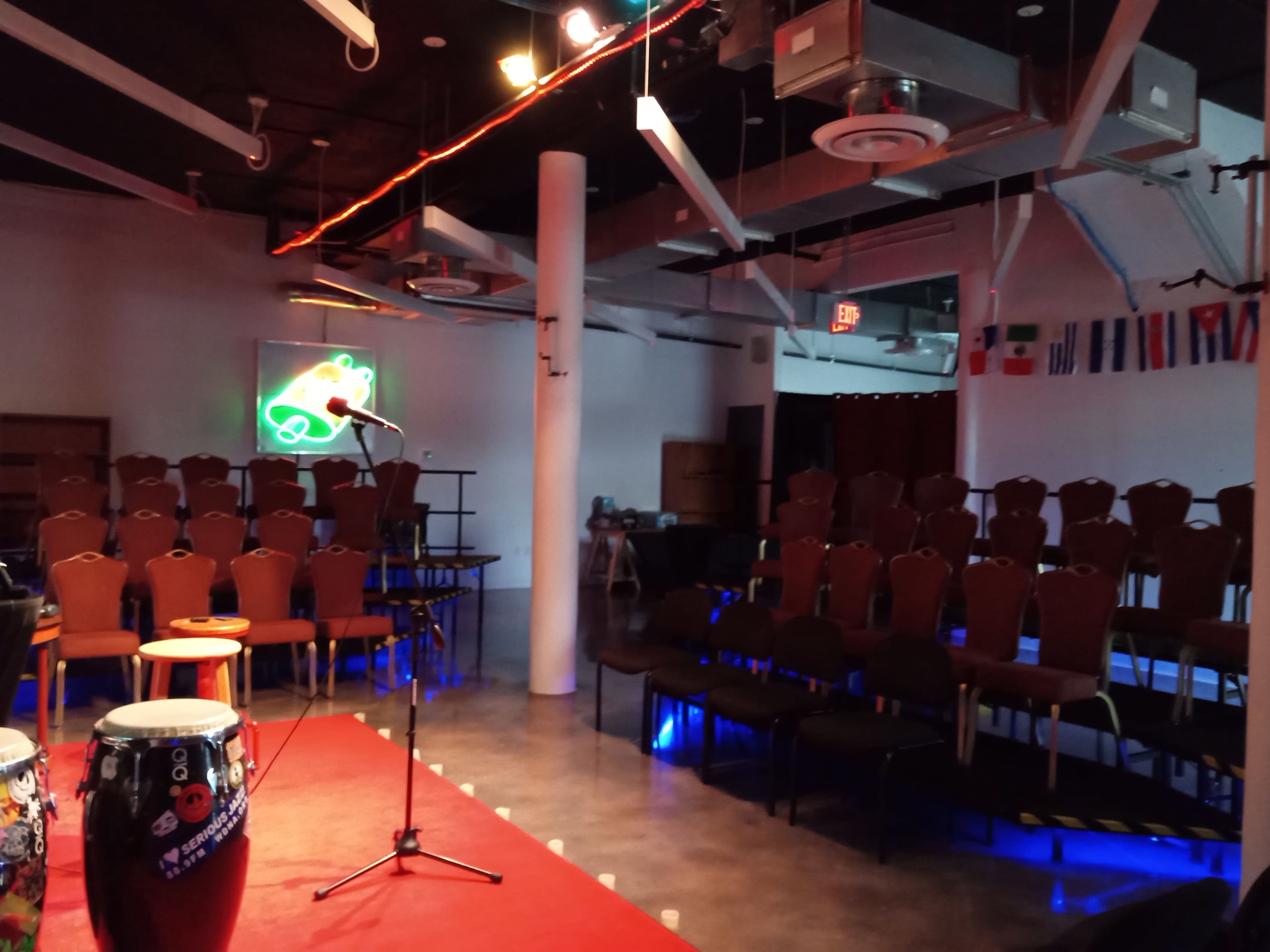 The image shows an indoor performance space with empty rows of chairs, a microphone on a small stage, and colorful decorative elements including flags and neon lights.