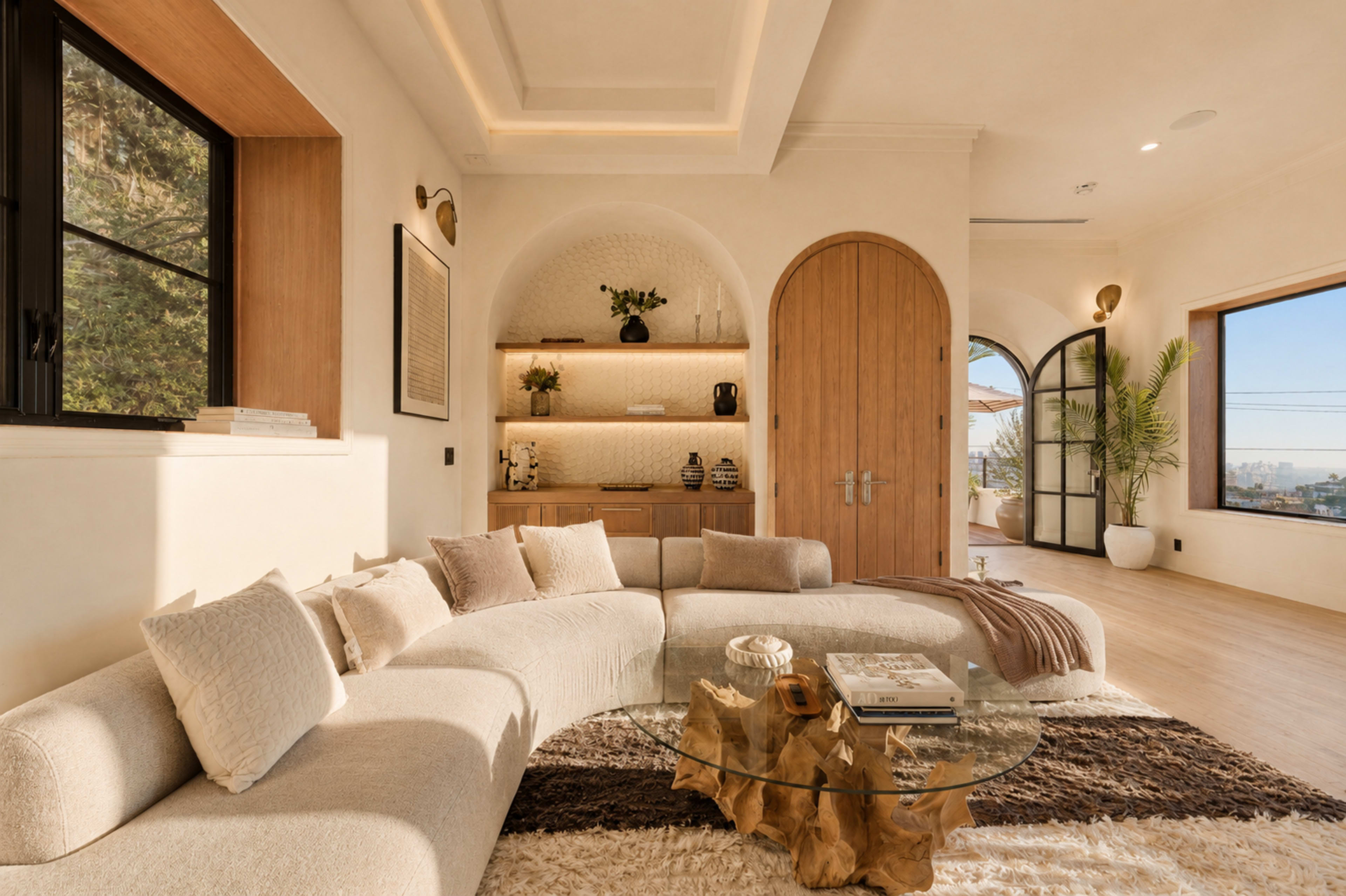 The scene features a modern living room with a curved beige sectional sofa, a round glass coffee table, and wooden accents, illuminated by natural light through large windows.