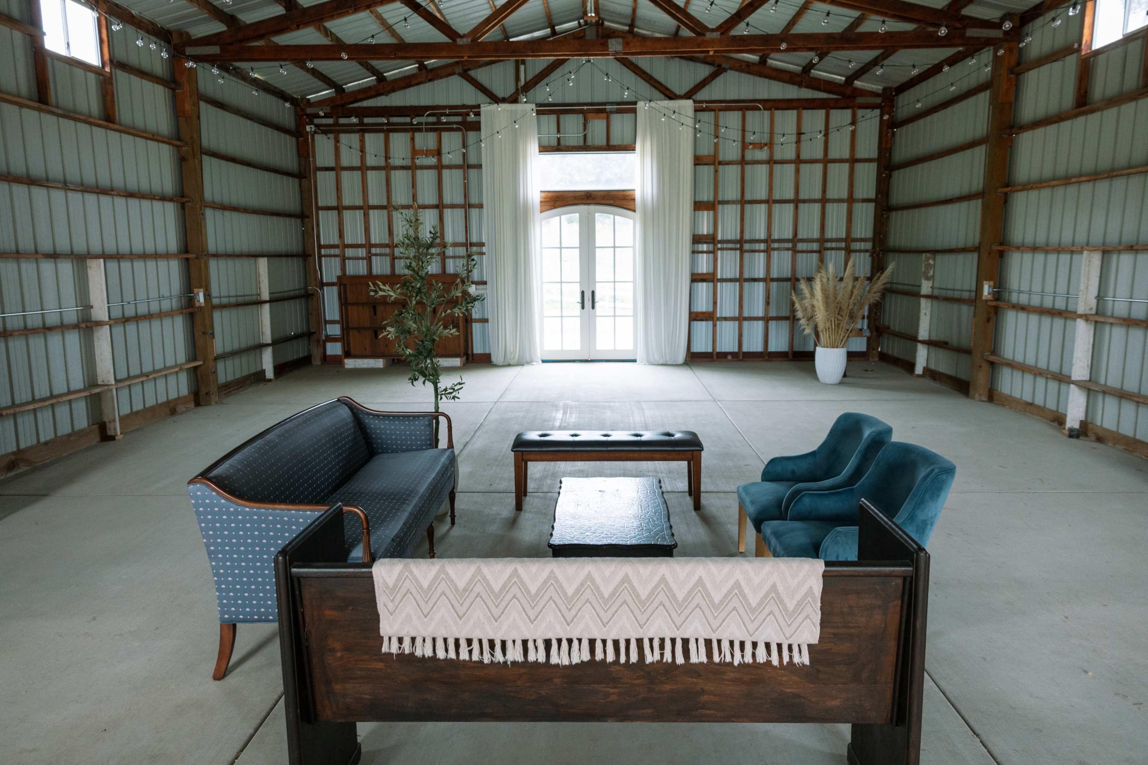 An empty interior space features a seating arrangement with a blue sofa, a wooden coffee table, and two chairs, surrounded by wooden beams and large glass doors.