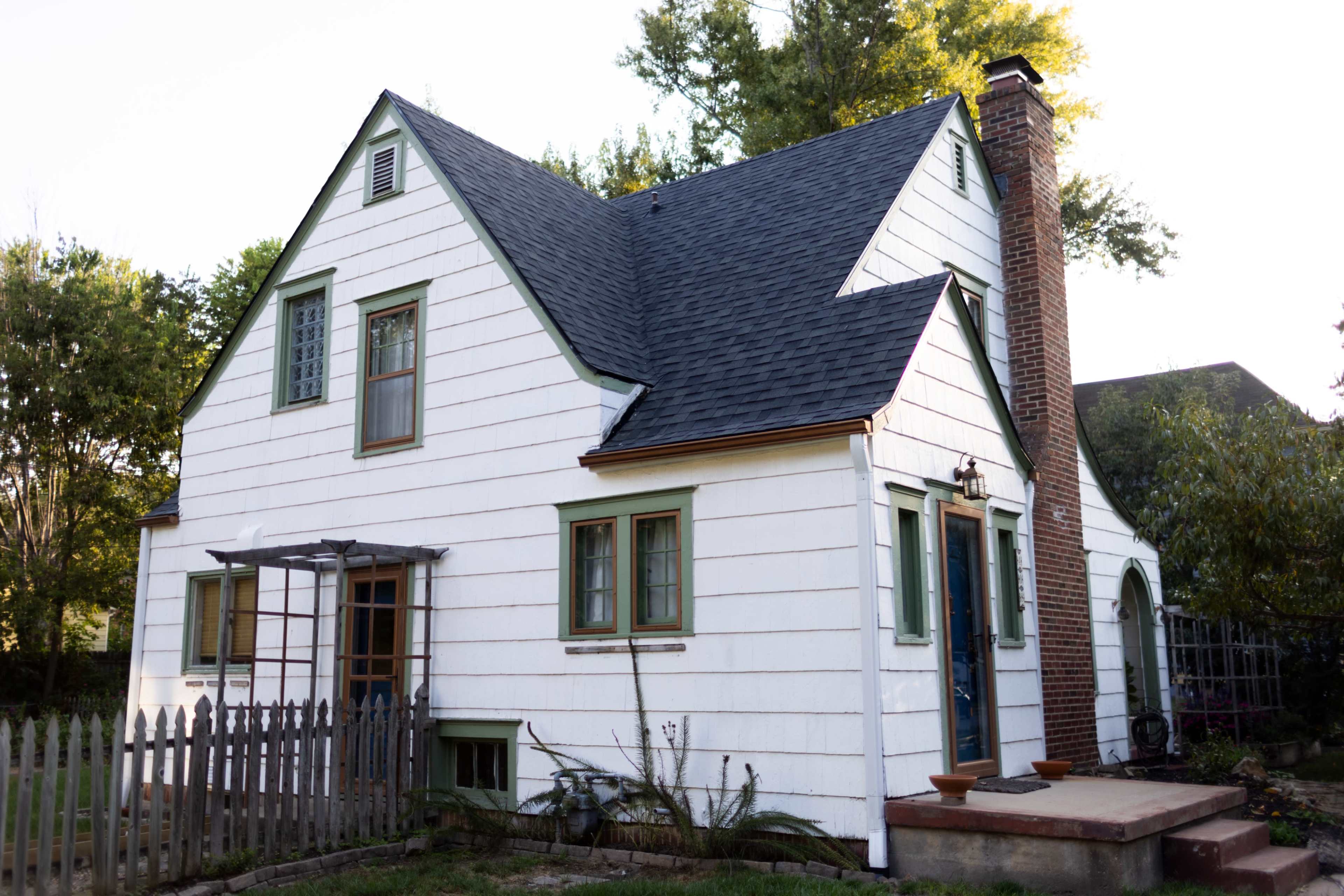 The image shows a two-story, white clapboard house with green trim, a steep roof, and a brick chimney surrounded by trees and a wooden fence.