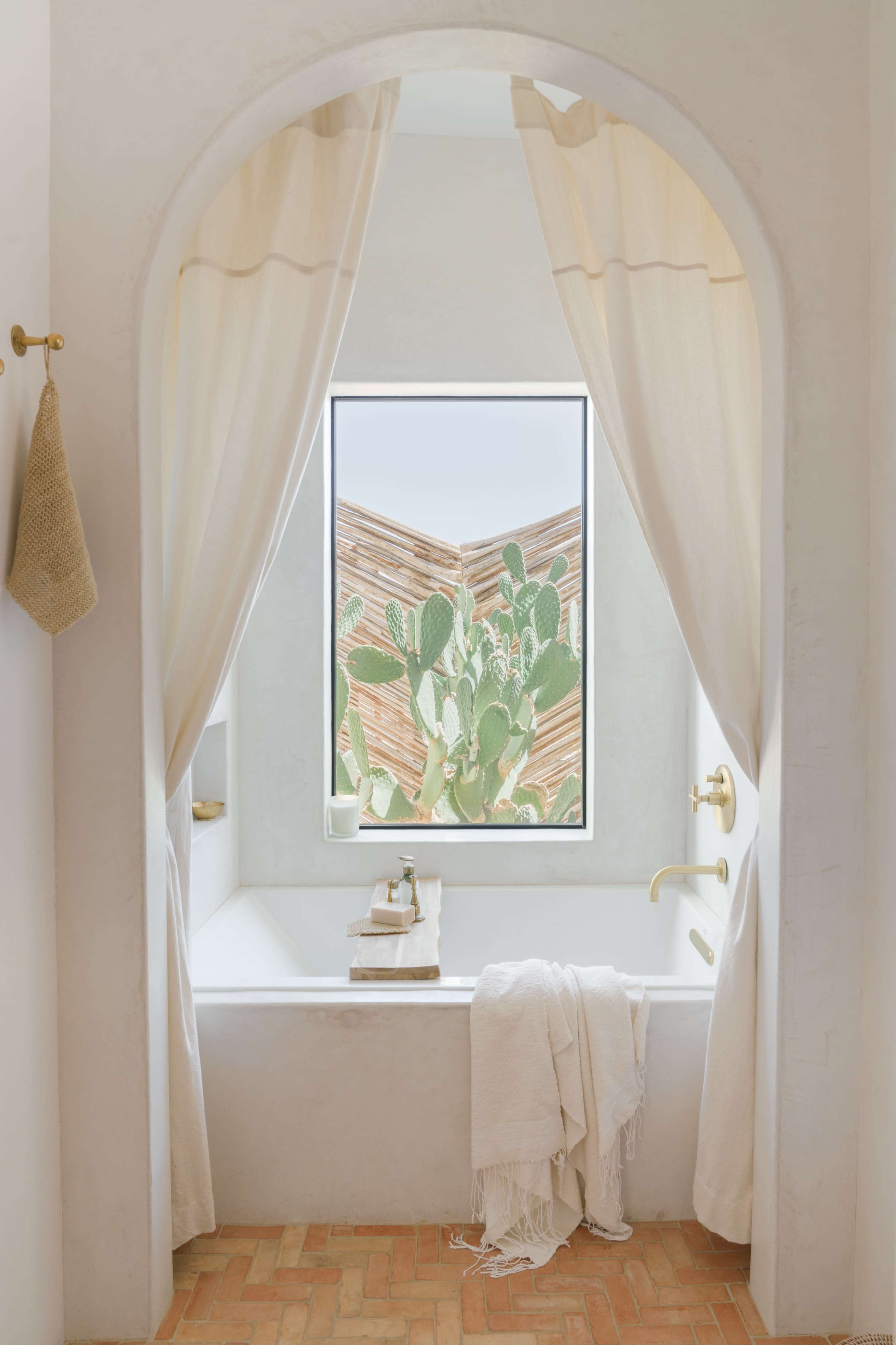 A bathtub is positioned under an archway, with sheer curtains framing a window that displays a view of a cactus garden outside.