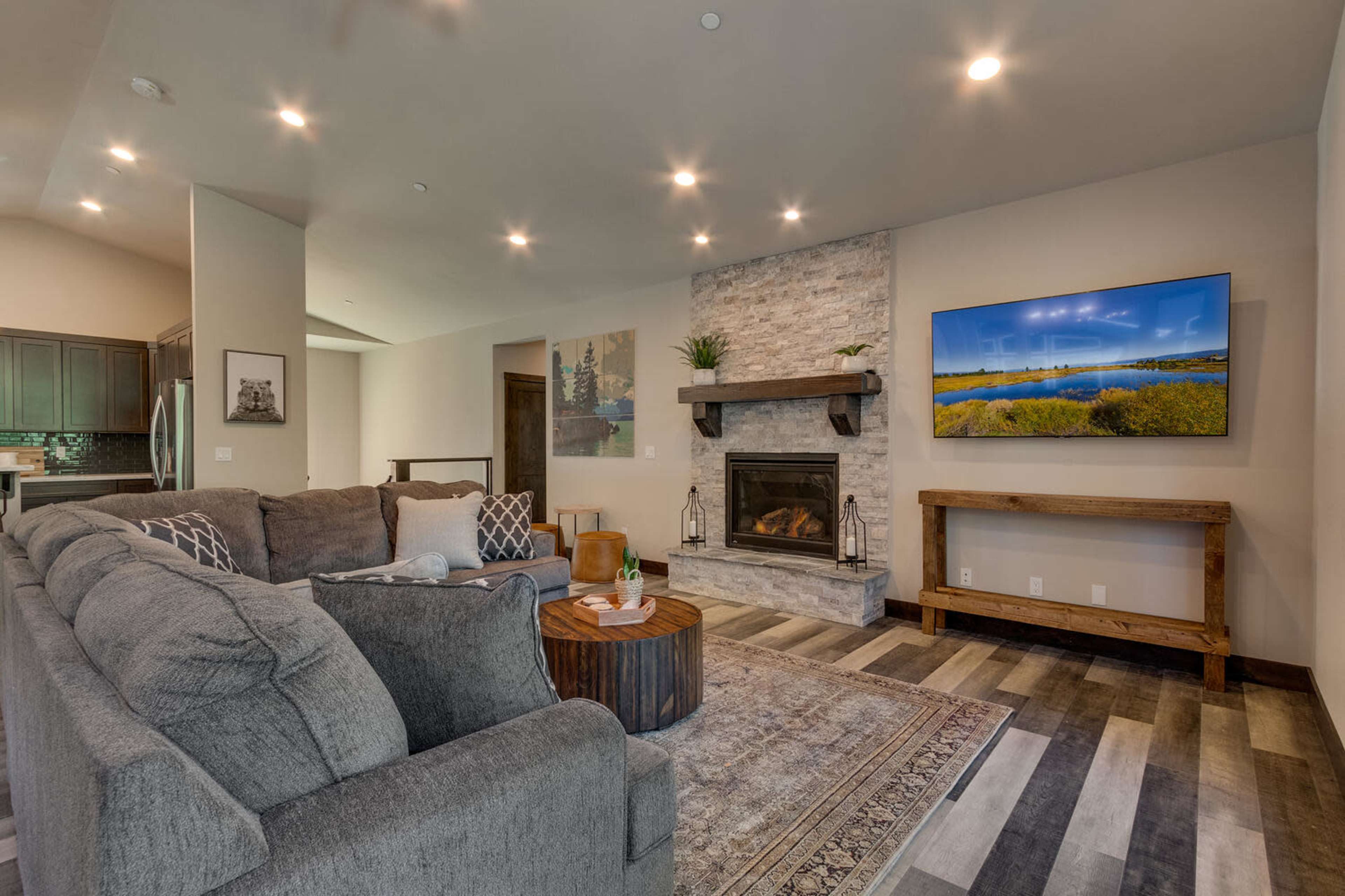 The living room features a stone fireplace, a wall-mounted TV, a grey sectional sofa, and a wooden coffee table on a patterned rug.