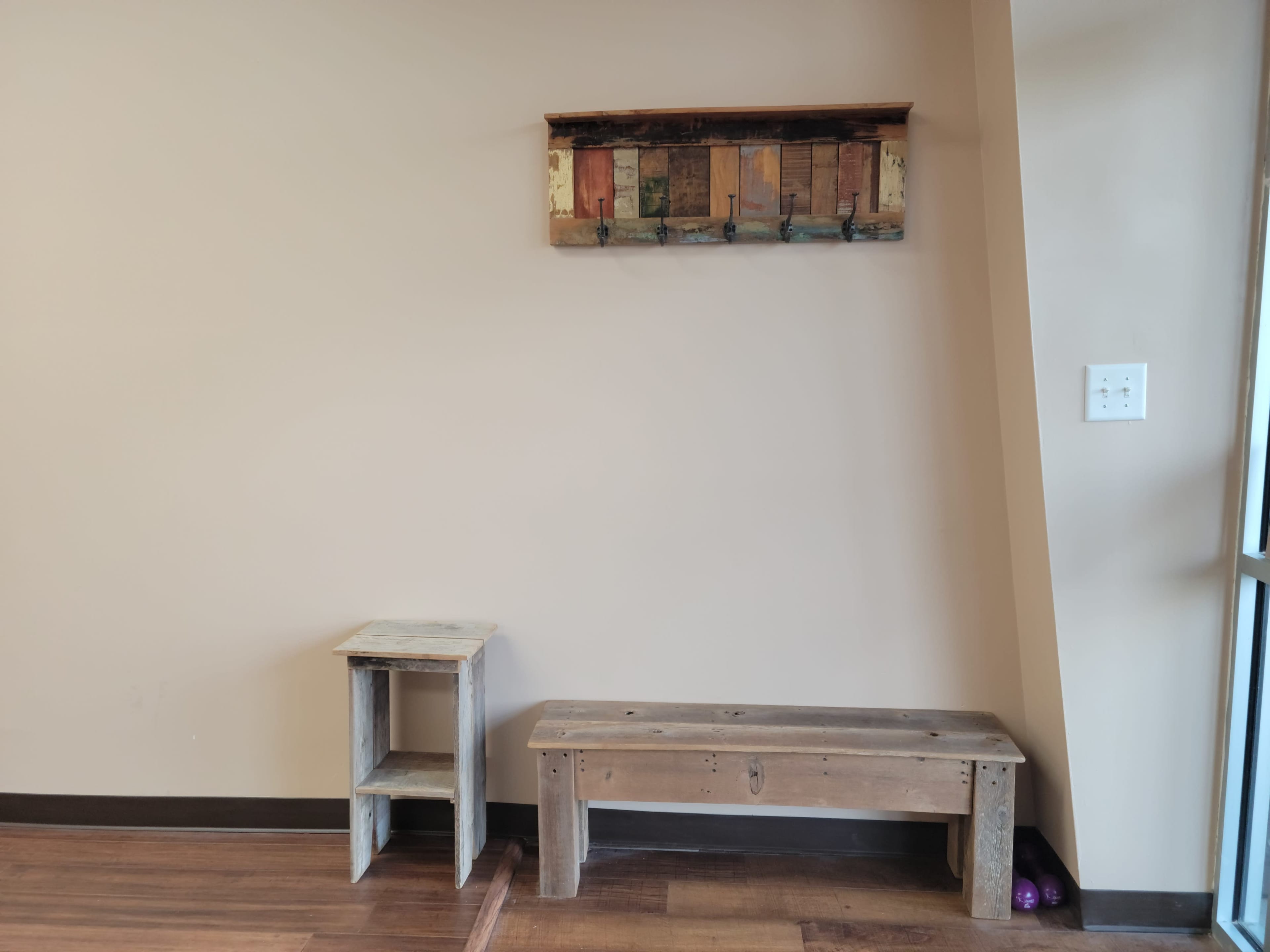 A simple interior wall with a wooden hook rack, a small side table, and a wooden bench on a hardwood floor.