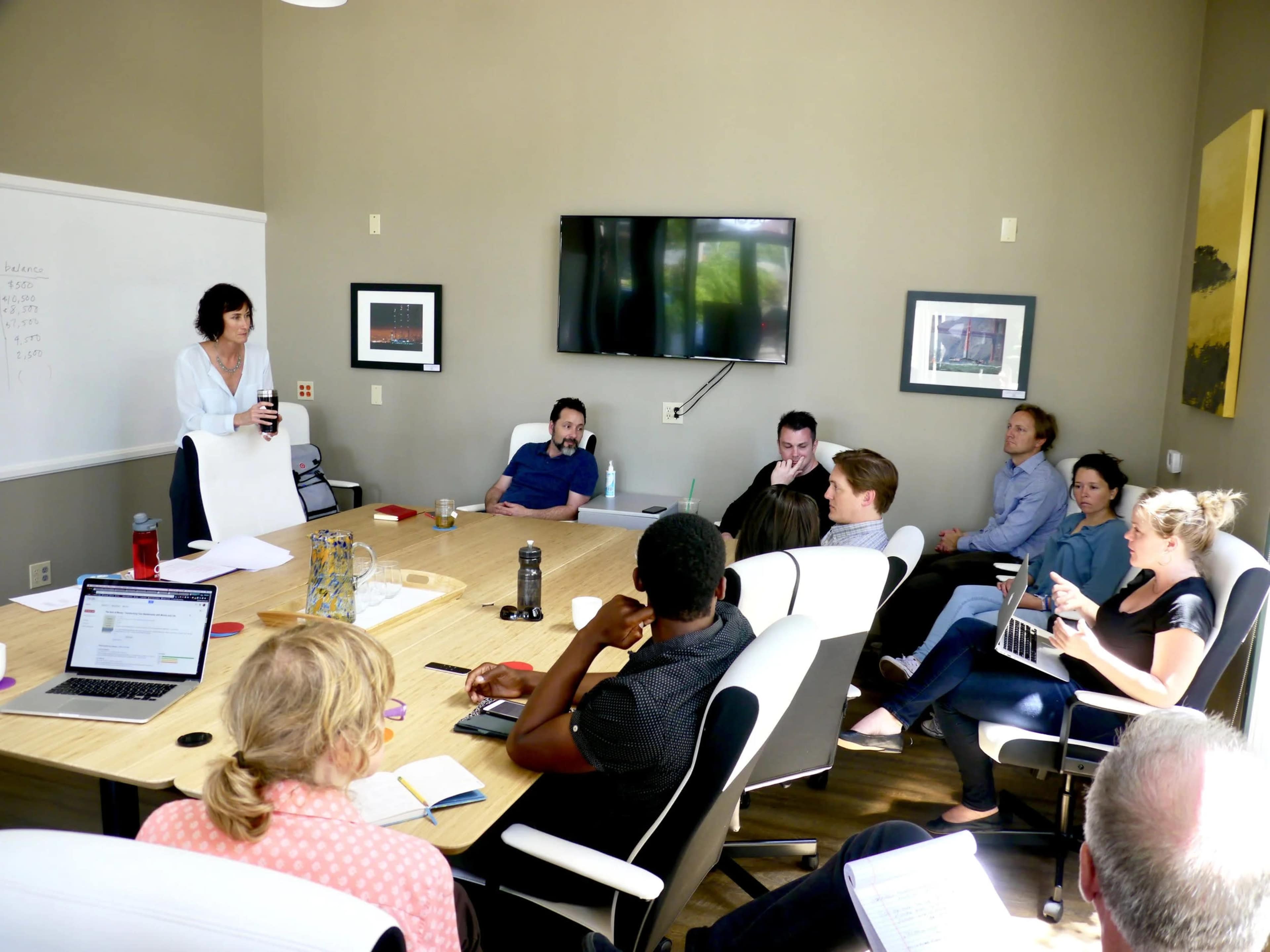 A group of people is sitting around a large conference table in a well-lit meeting room, while one person stands at the front presenting.