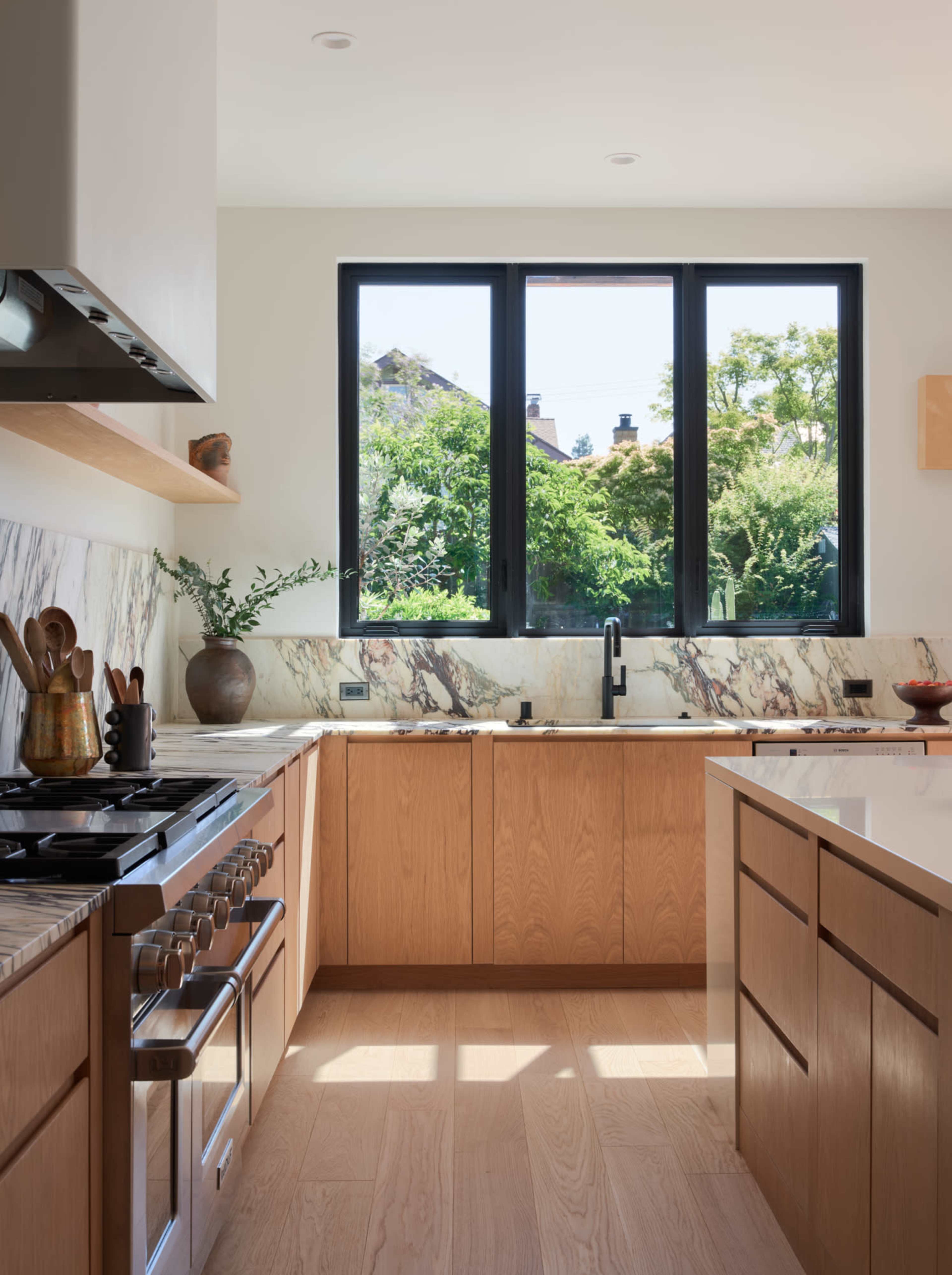 The kitchen features wooden cabinetry, a marble countertop, a gas stove, and large windows that provide a view of greenery outside.