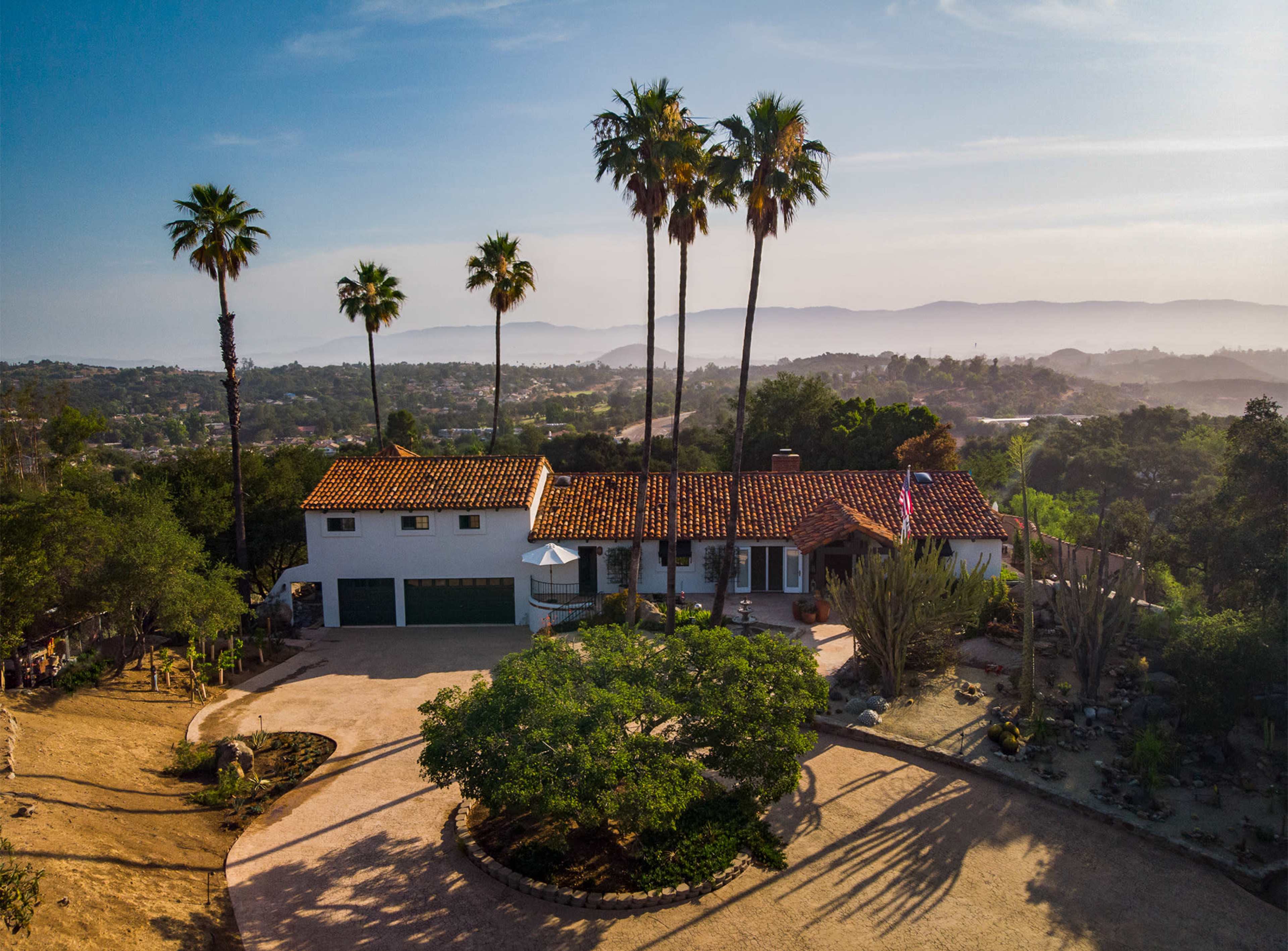A large, two-story house with a tile roof is surrounded by palm trees and desert landscaping, located on a hillside with a view of distant mountains.