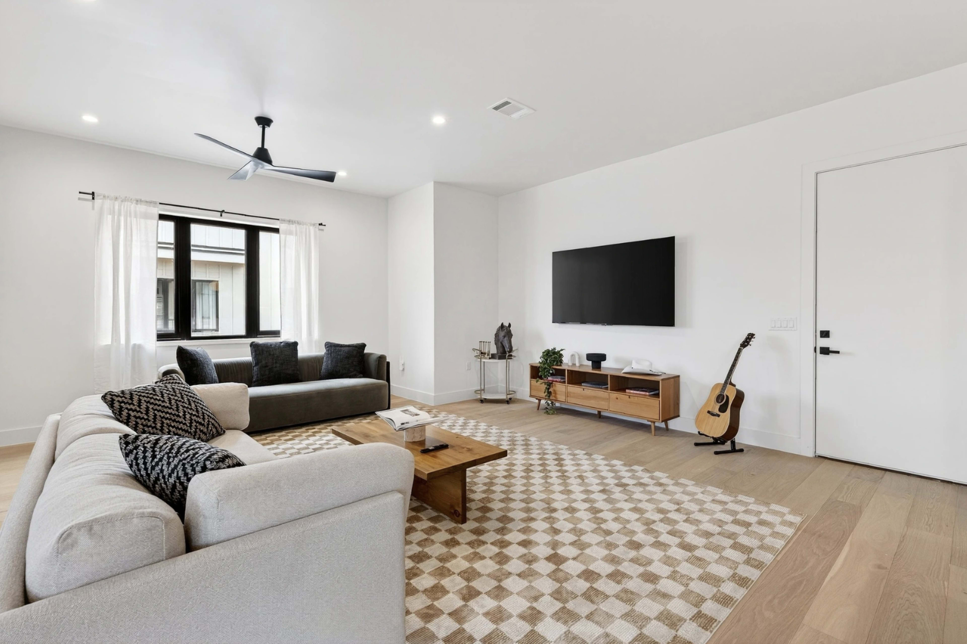The living room features a sofa, a coffee table, a guitar, and a mounted television on the wall, with natural light coming through a window.
