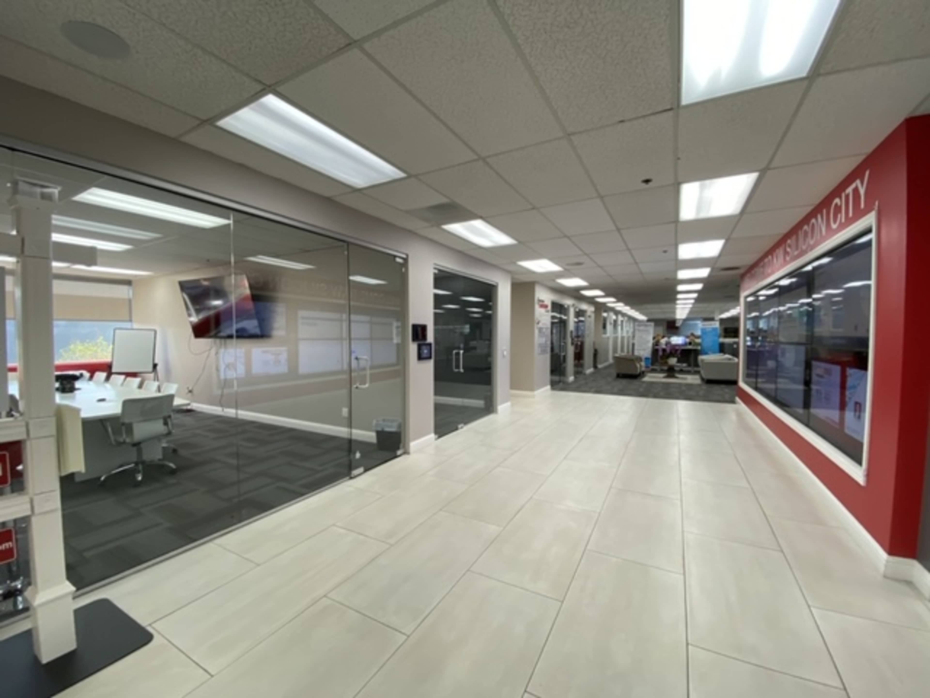 The image shows a modern office interior with glass conference rooms, white tiled flooring, and a red accent wall displaying a promotional panel.