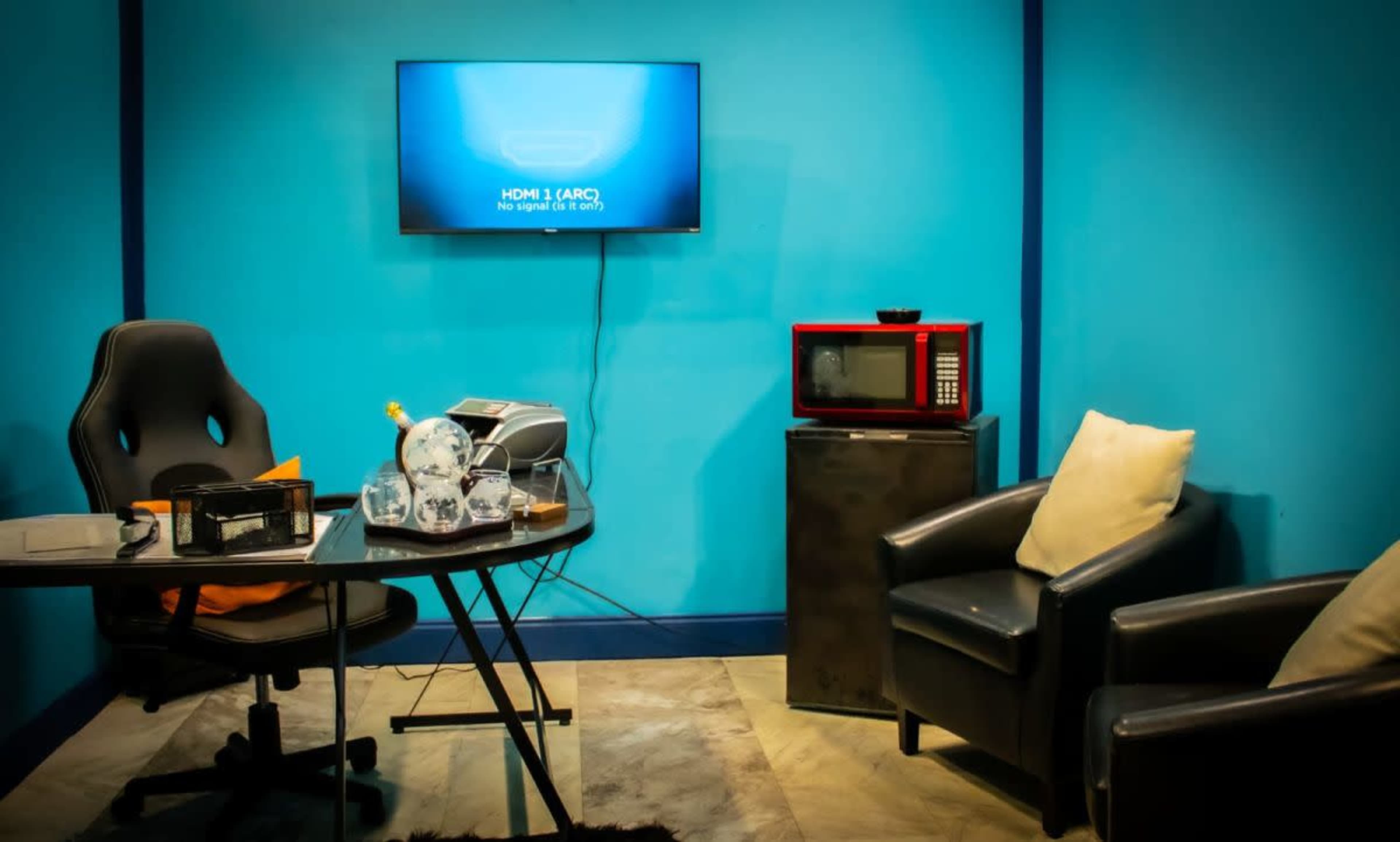 The room features a bright blue wall, a television displaying a "No Signal" message, a black office chair, a table with a printer, a microwave, and a small black cabinet beside two cushions.