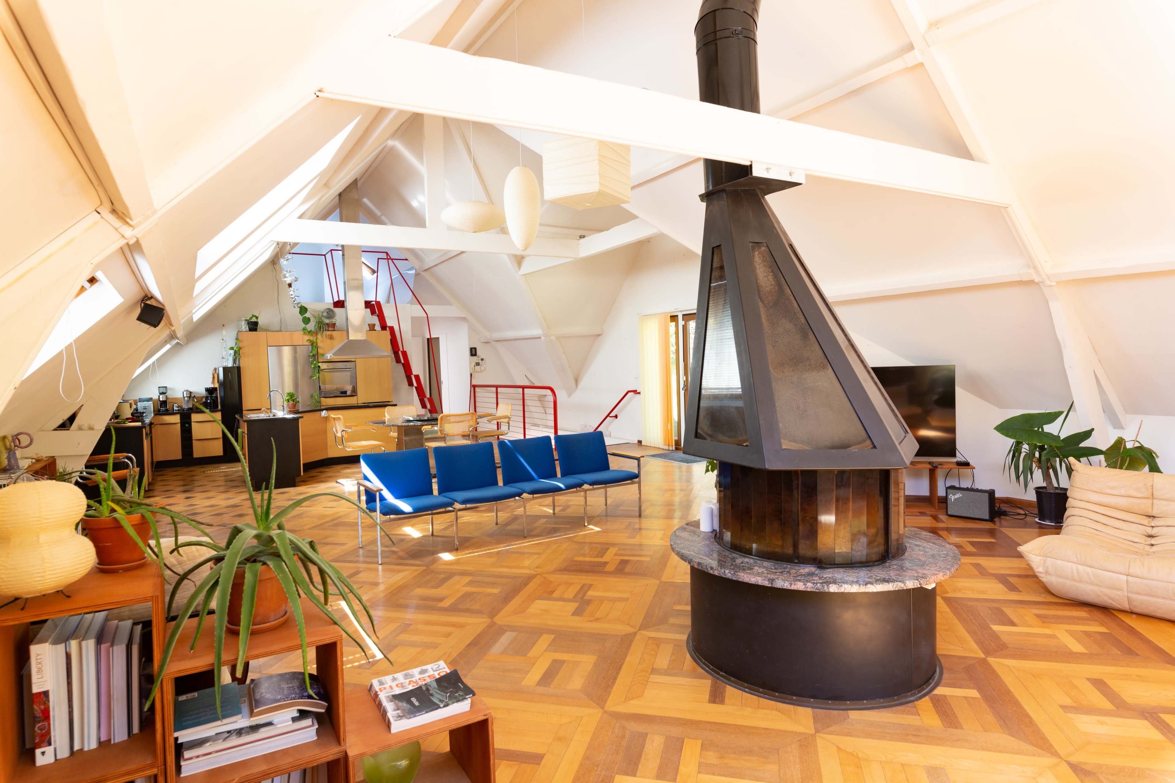 The image shows a spacious, modern living area with a central fireplace, wooden flooring, and large windows.