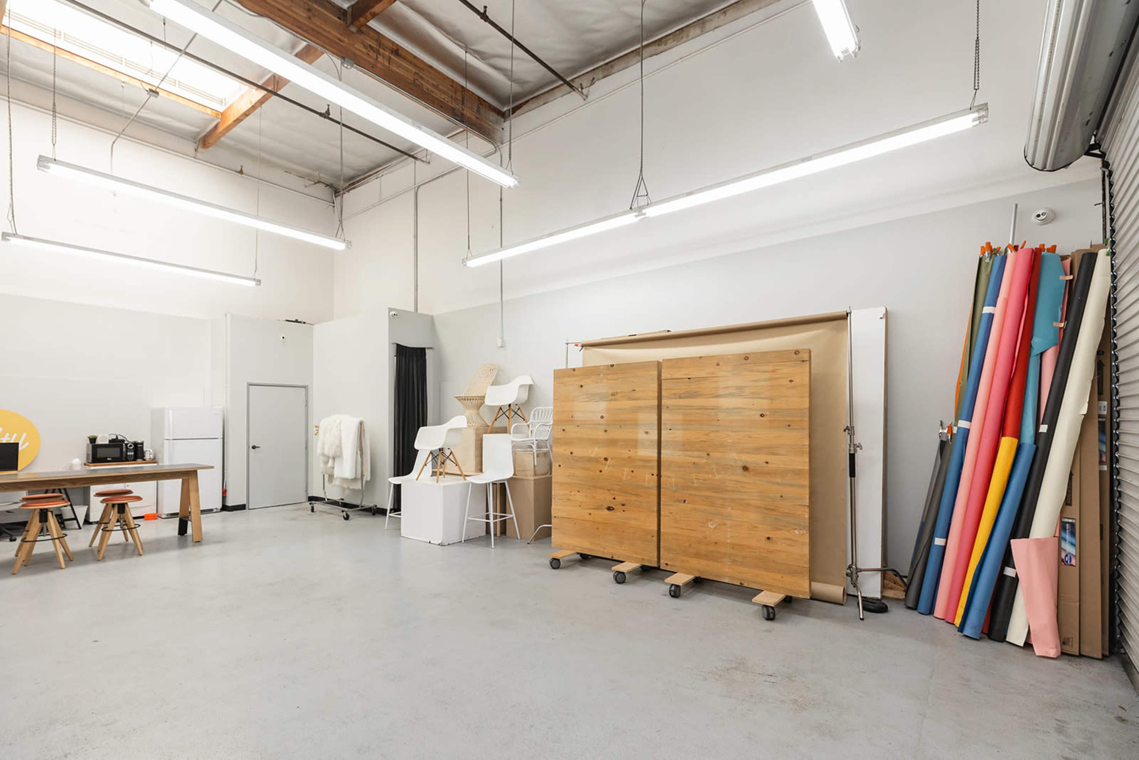 A spacious studio with a wooden storage unit, a small table with stools, and several rolls of colorful material stacked against the wall.