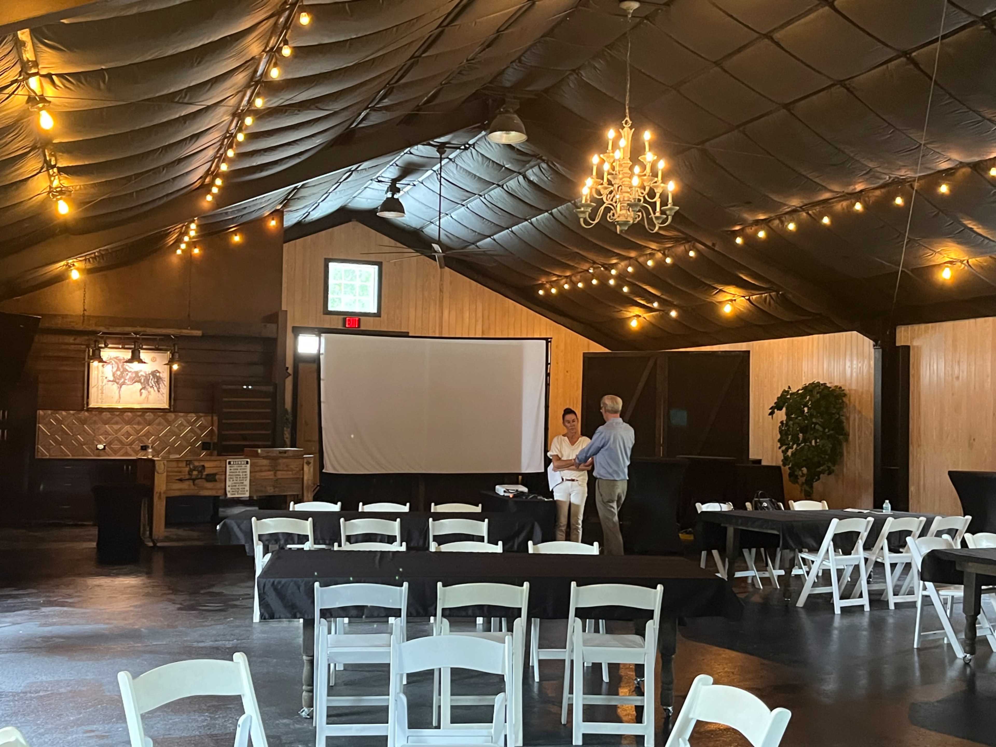 A large indoor venue with wooden walls, a high ceiling, and string lights is set up with black tables and chairs, featuring a projector screen at the front.