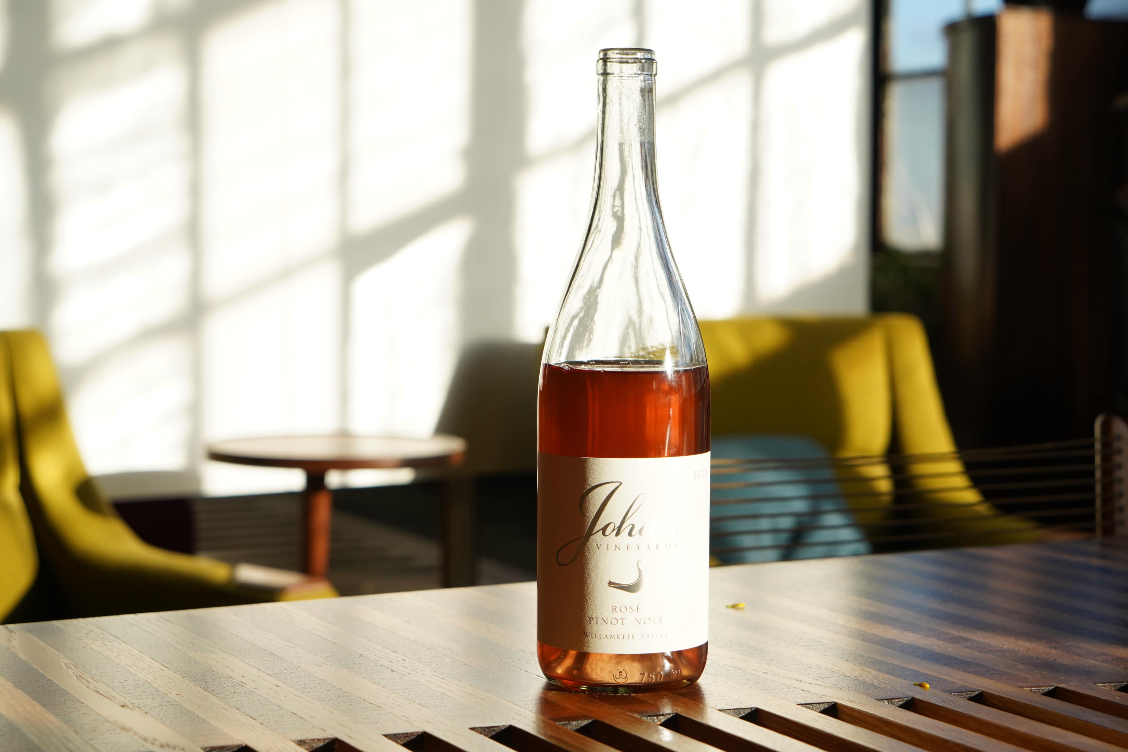 A bottle of rosé wine is placed on a wooden table, illuminated by sunlight, with two green chairs visible in the background.