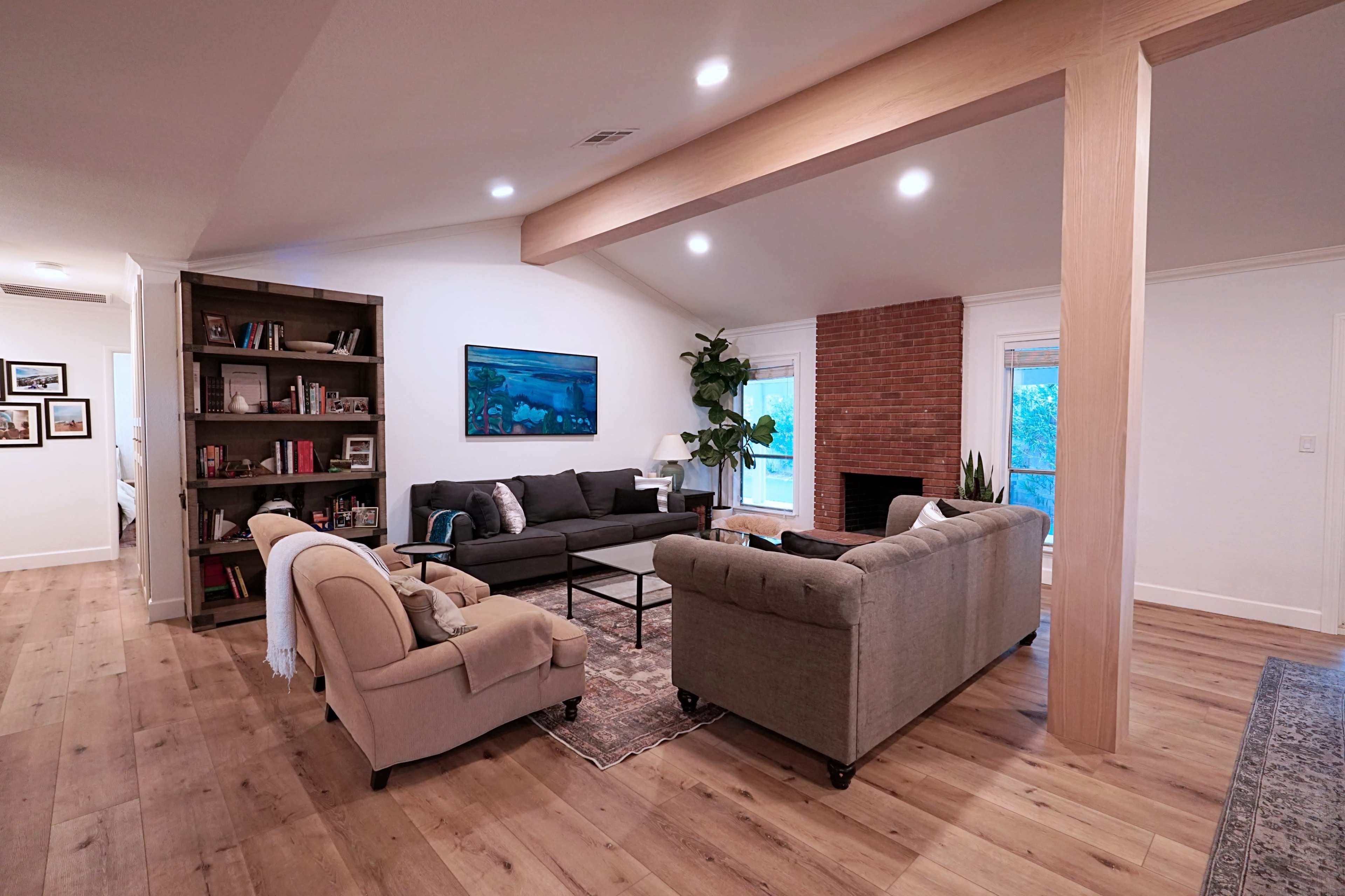 A modern living room featuring two sofas, a bookshelf, a brick fireplace, and large windows.