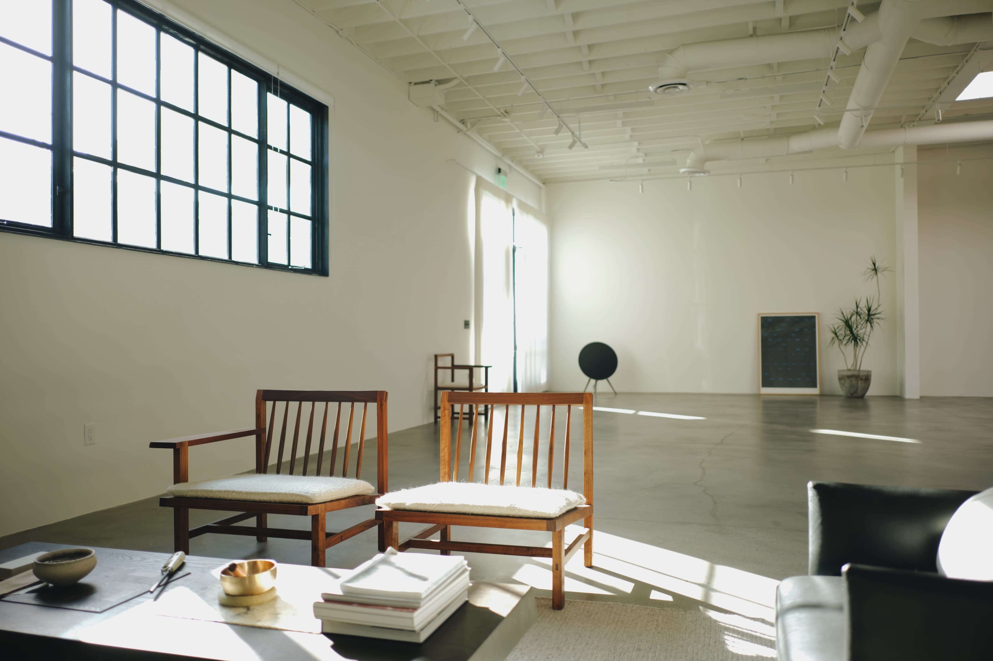 The image shows a minimalist interior space with two wooden chairs, a coffee table, and a large empty area illuminated by natural light.