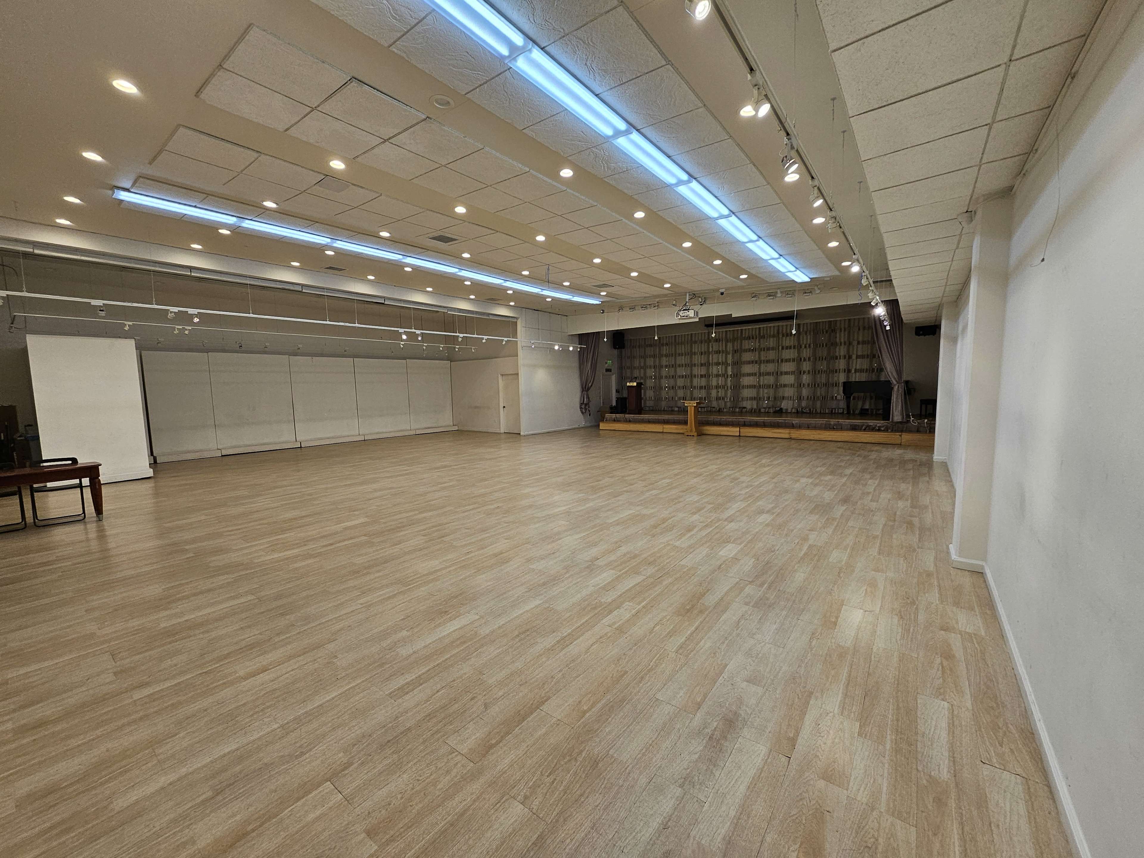 A spacious, well-lit hall with wooden flooring, featuring a stage at the far end and a few tables arranged on one side.