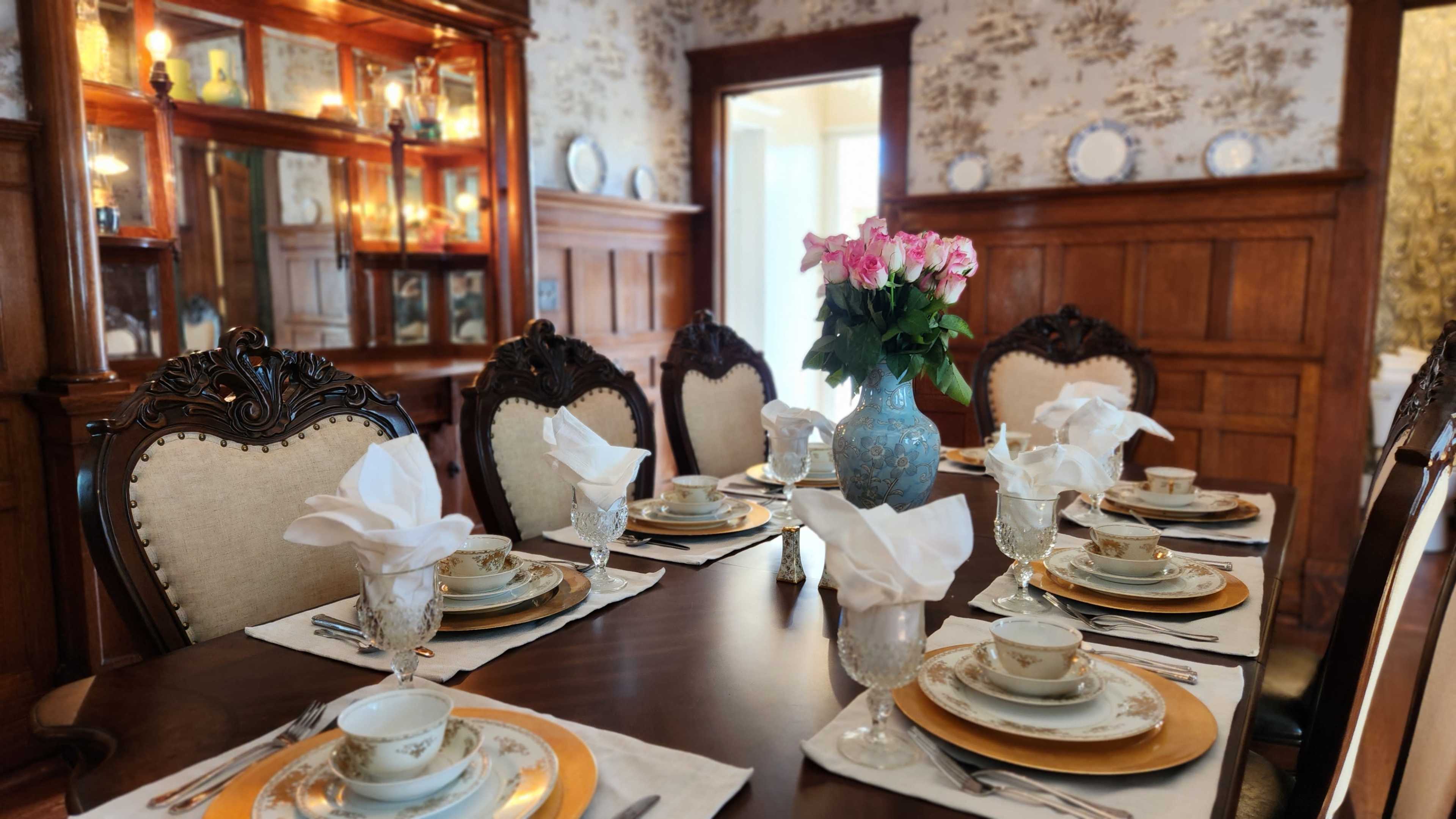 A dining table is set with fine china, glassware, and a vase of roses, surrounded by elegantly upholstered chairs in a room with wooden paneling and ornate decorations.