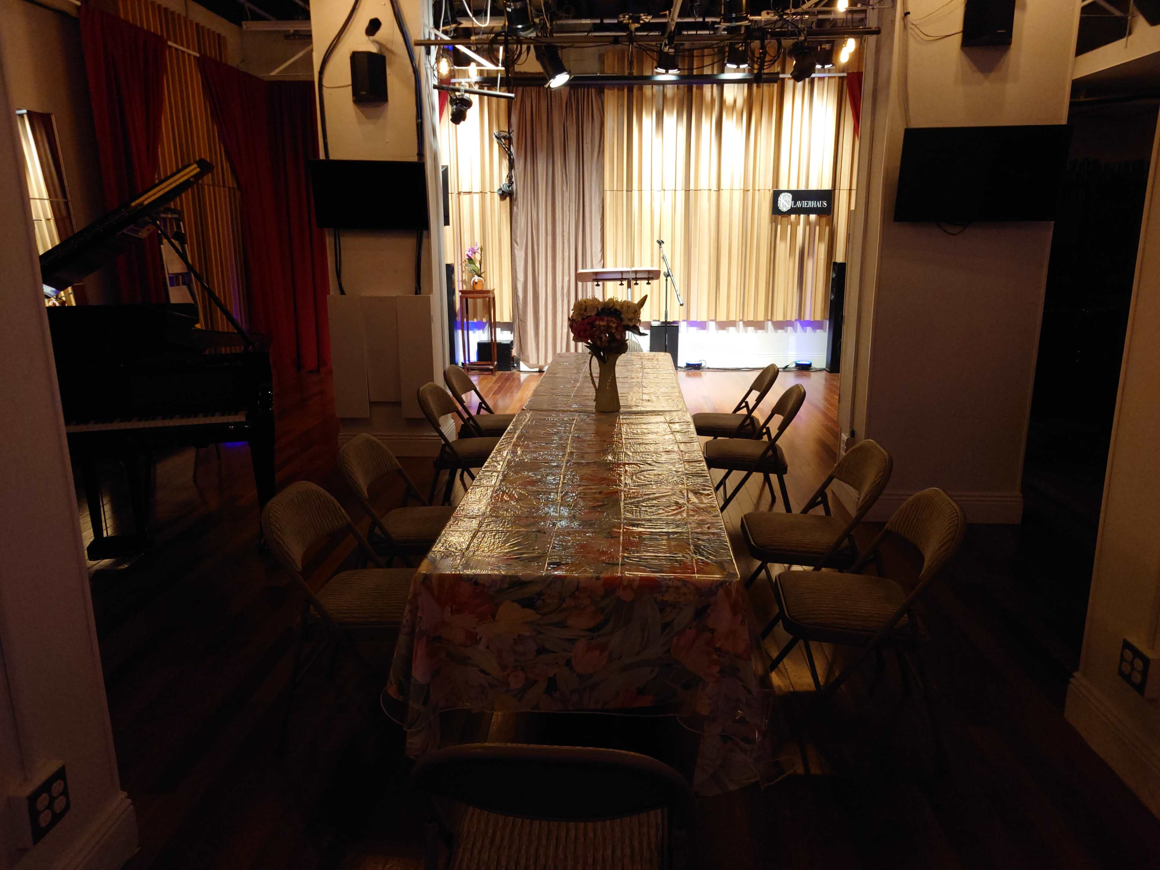 A large, rectangular table with chairs is set in a well-lit room featuring a stage and a grand piano.