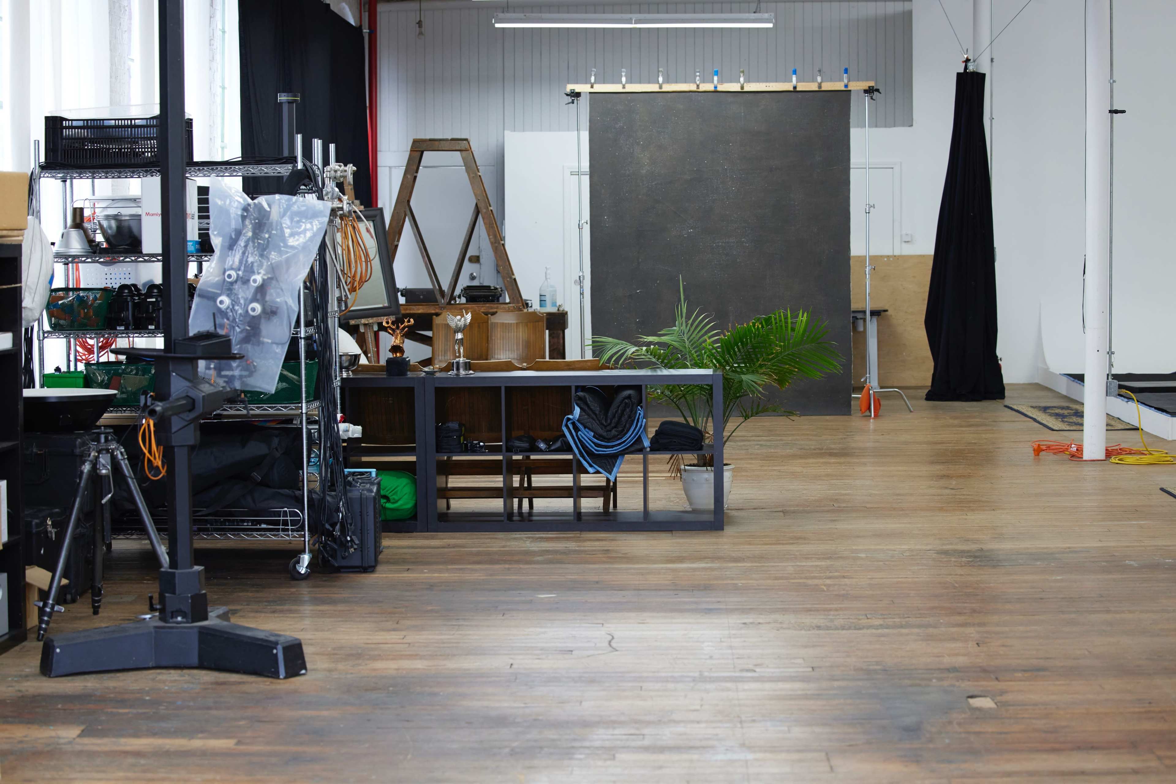 A spacious studio with wooden flooring, various furniture and equipment, including a backdrop, shelves, and a table, arranged in an organized manner.