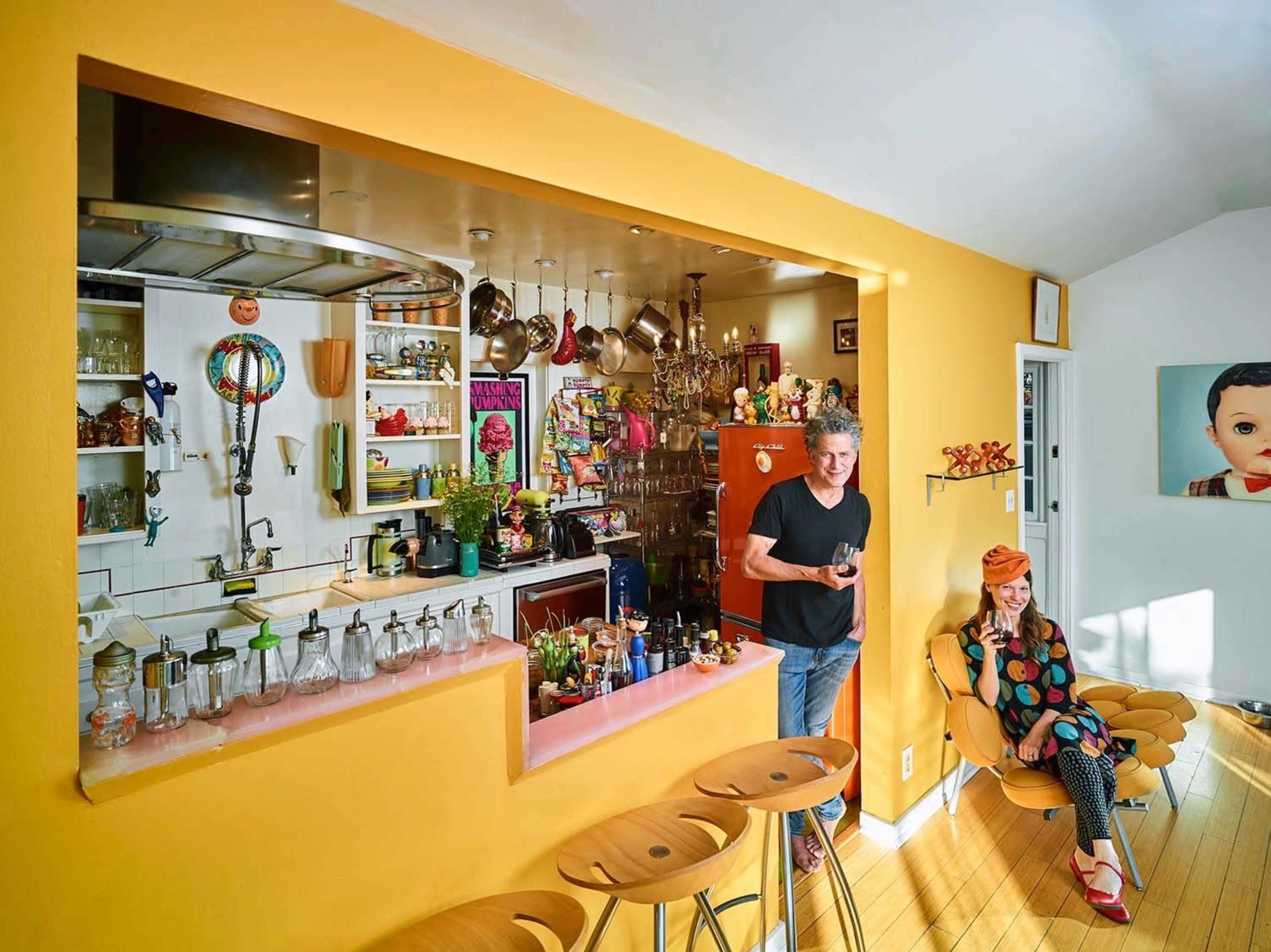 A brightly colored kitchen features a man standing by the counter and a woman sitting in a chair, surrounded by various kitchen items and decorations.