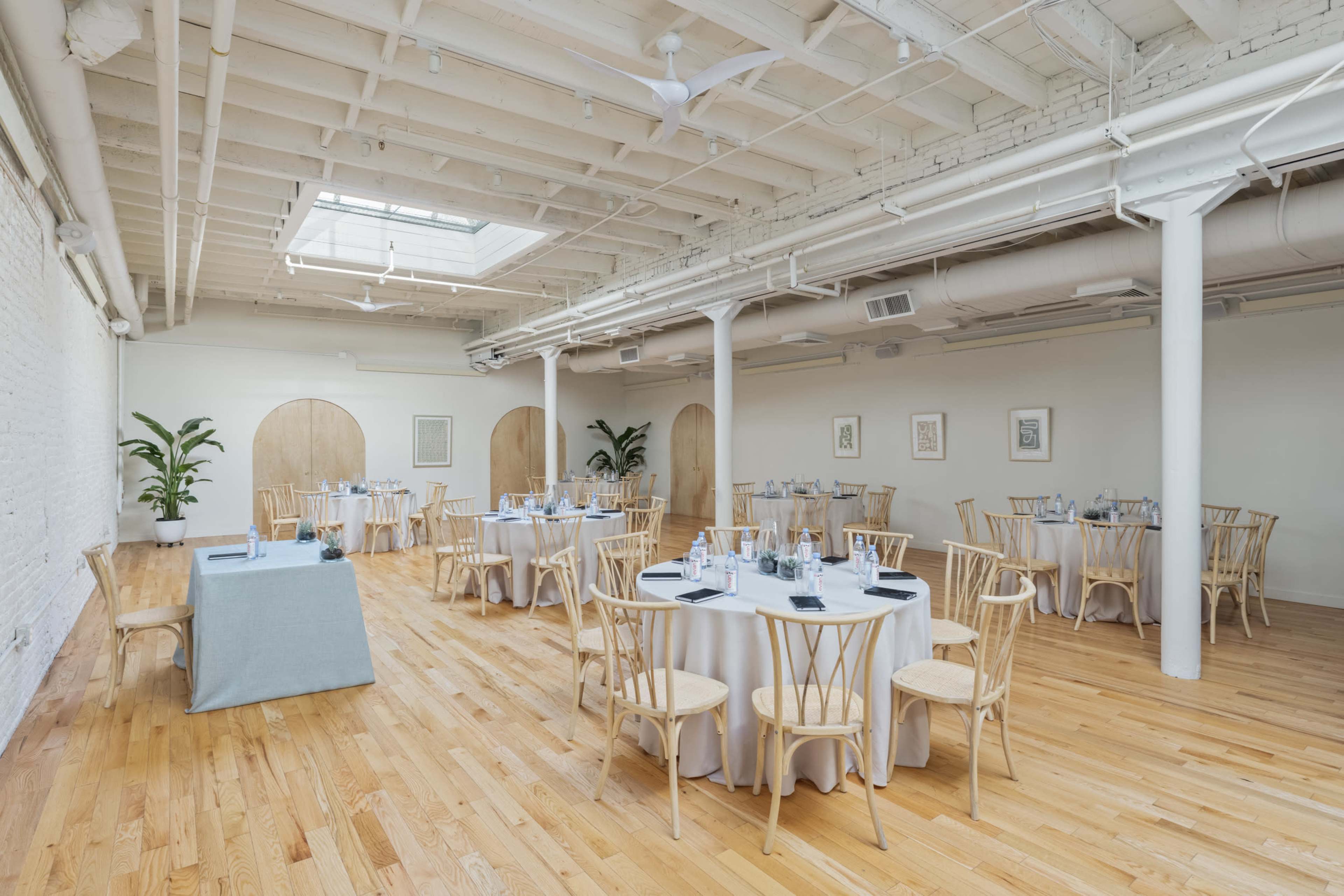 A spacious event room with round tables set for dining, hardwood floors, and large windows allowing natural light to enter.