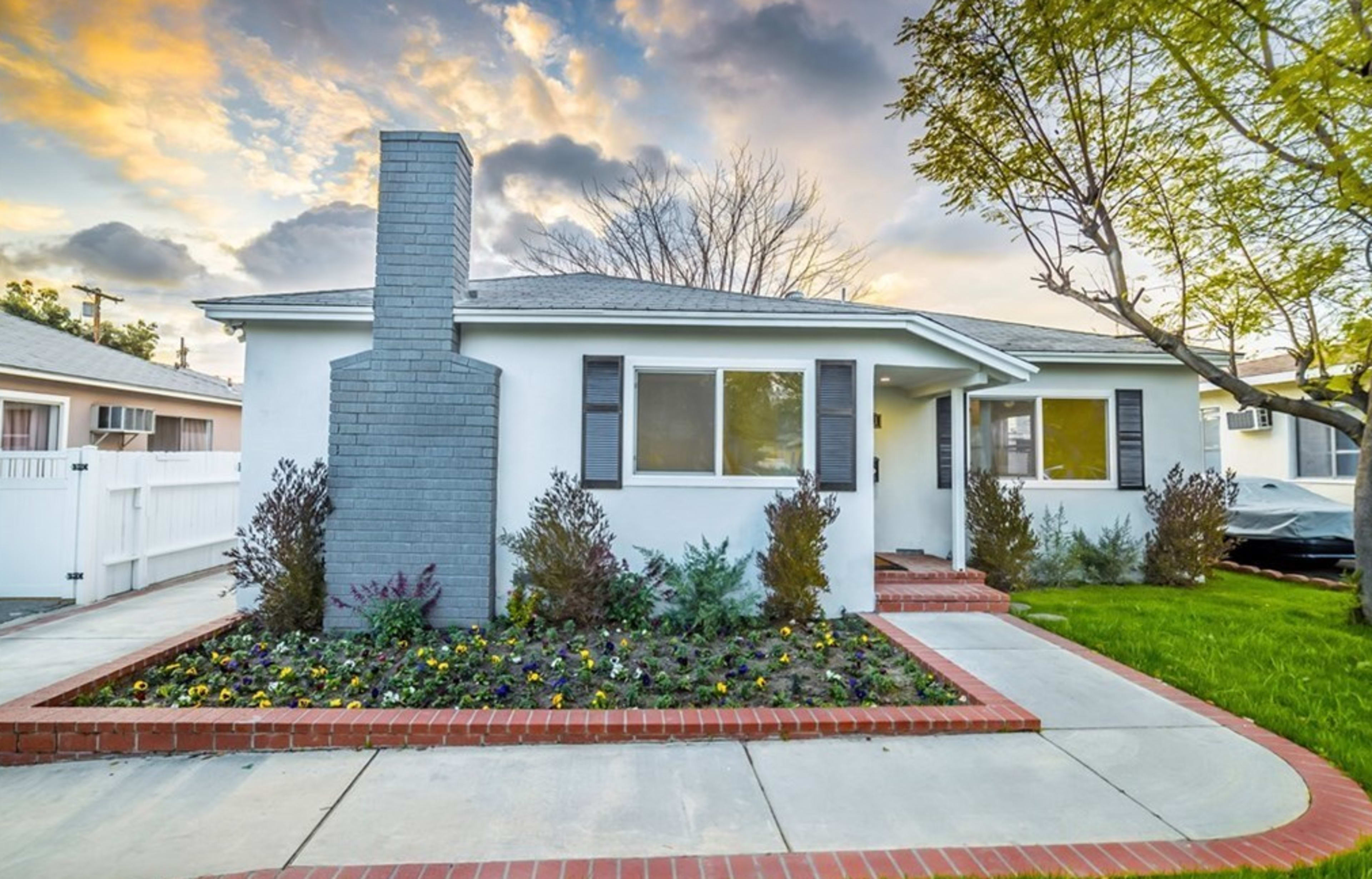 A single-story house with a gray chimney, surrounded by a landscaped front yard featuring colorful flowers and a paved walkway.