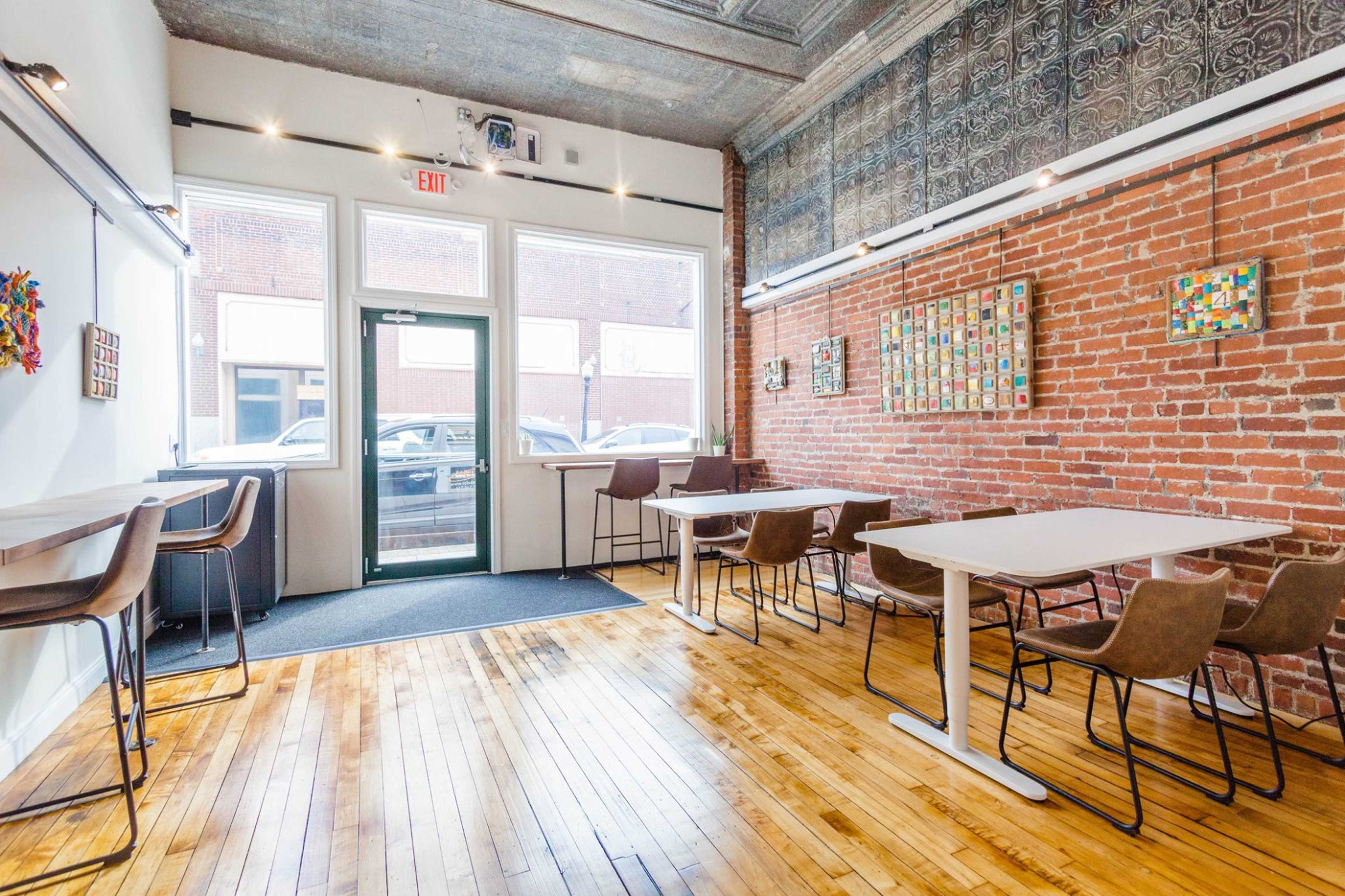 A bright, modern café features wooden floors, exposed brick walls, and various seating arrangements with tables and chairs near large windows.