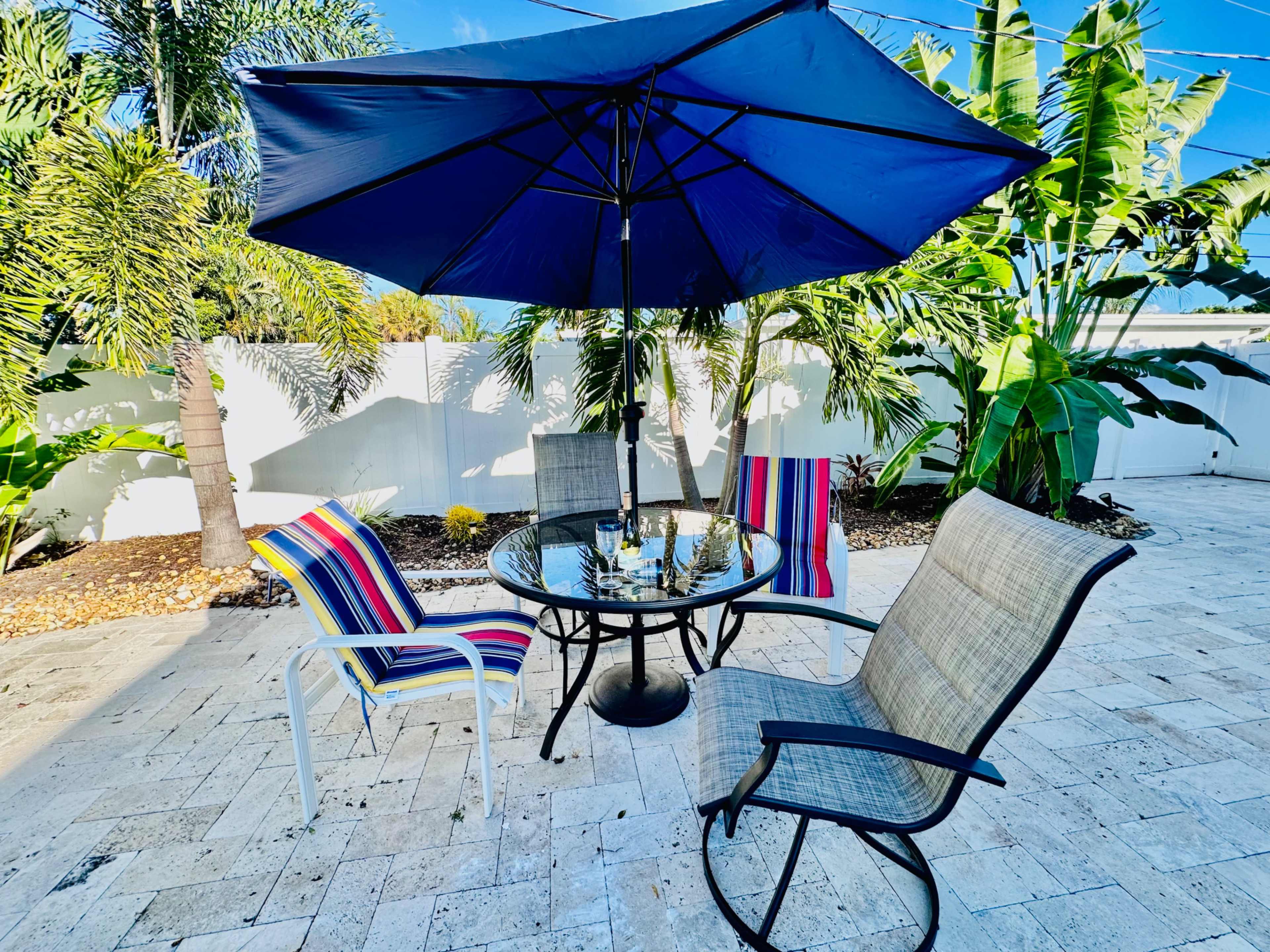 A patio area features a round table with four chairs under a large blue umbrella, surrounded by tropical plants.