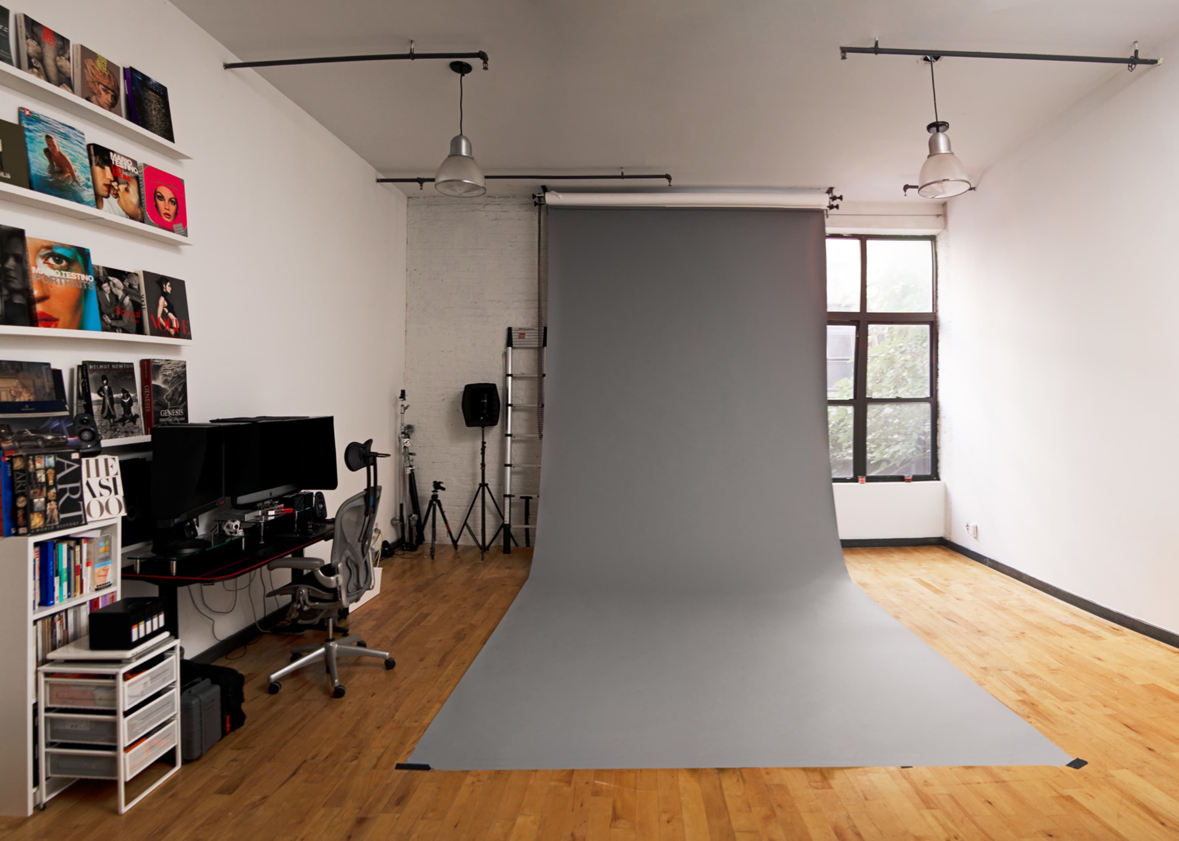 The image shows a minimalist studio space with a gray photography backdrop, a desk with dual monitors, and various equipment arranged along the walls.
