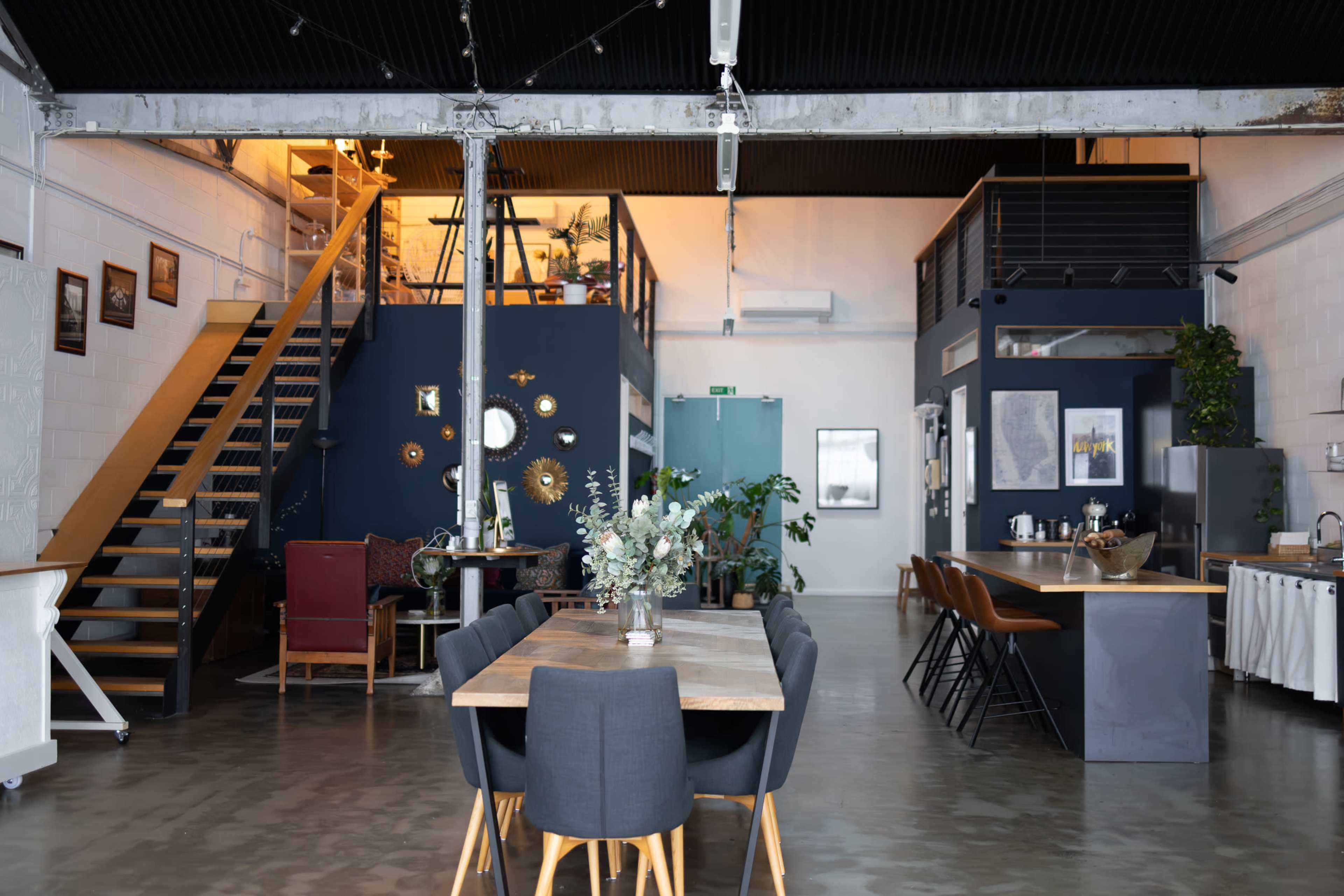 The image shows an industrial-style open space featuring a dining table surrounded by chairs, a kitchen area, and a staircase leading to a lofted section.