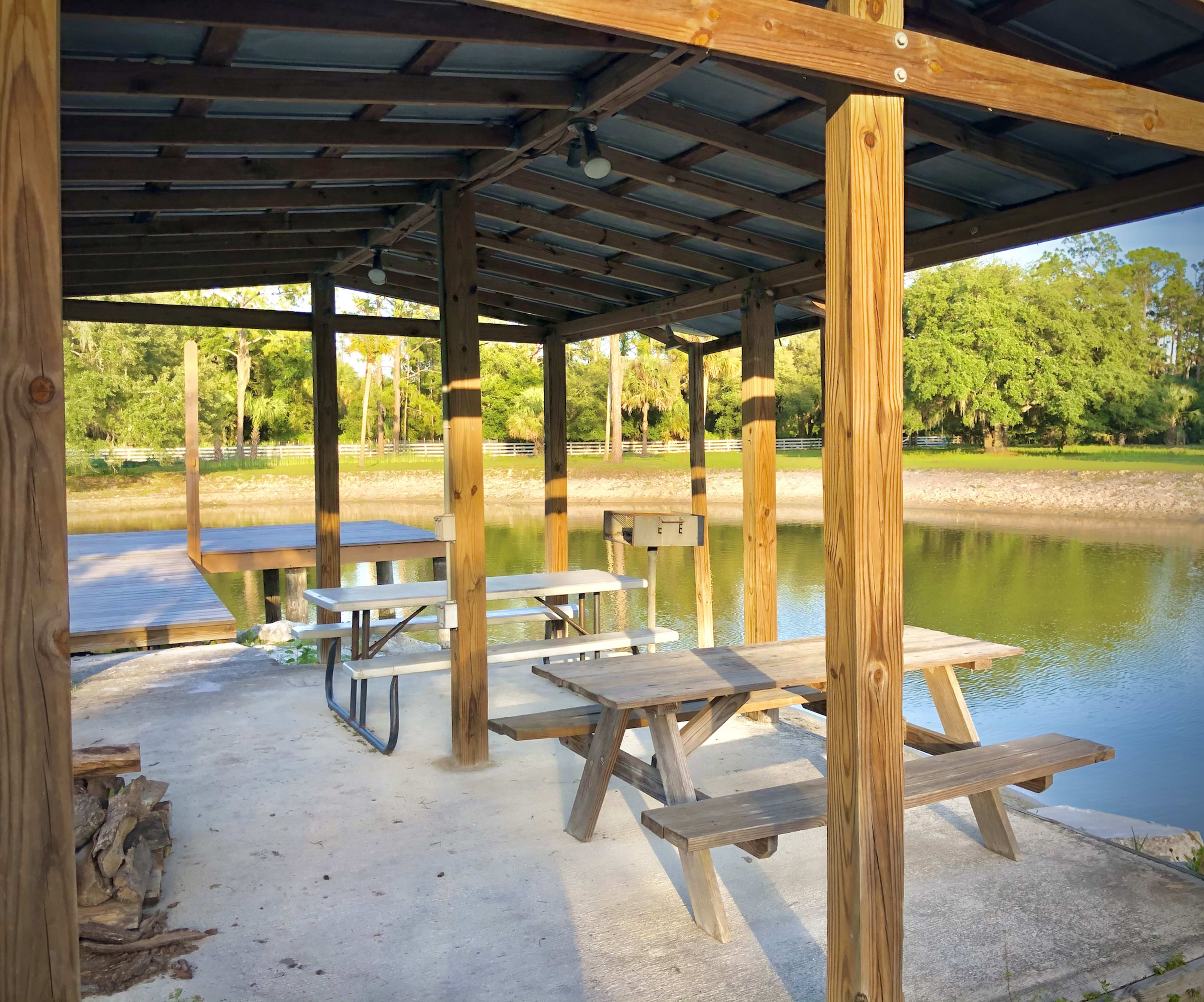 The image shows a wooden shelter with picnic tables by a calm body of water, surrounded by trees.