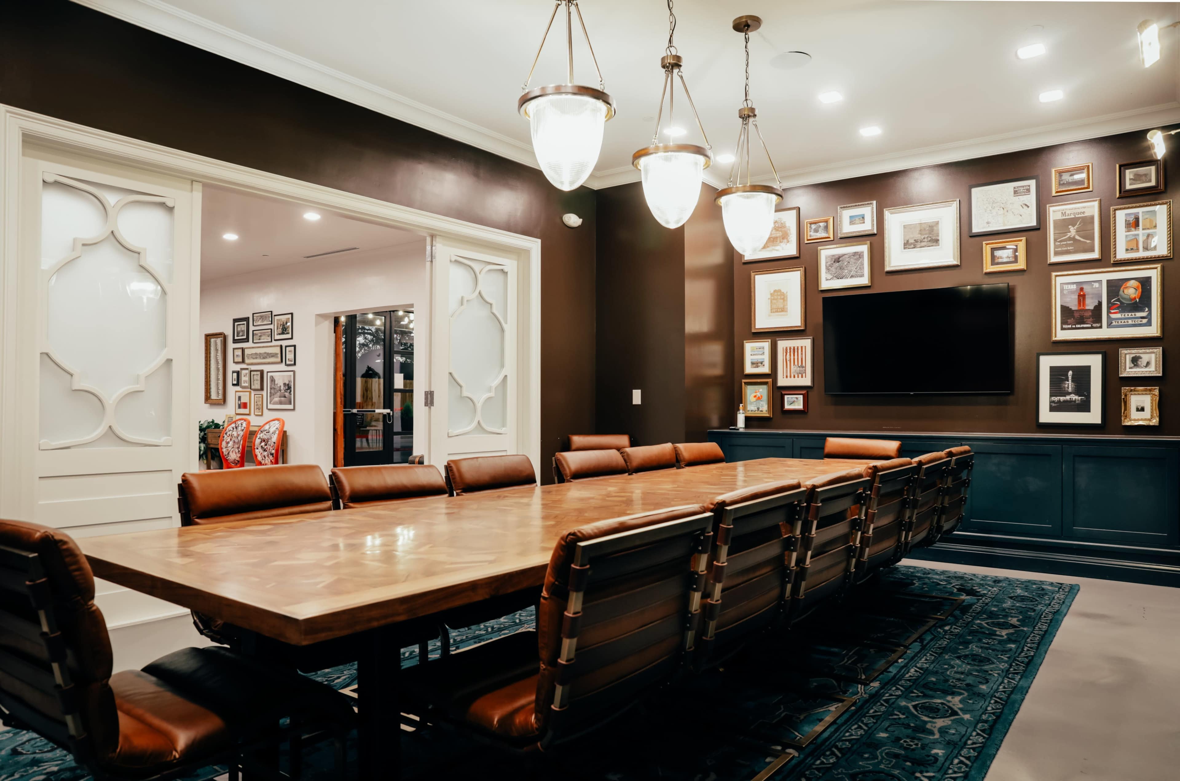 A large conference room features a wooden table surrounded by leather chairs and a wall adorned with framed artwork, with a television mounted above a dark cabinet.