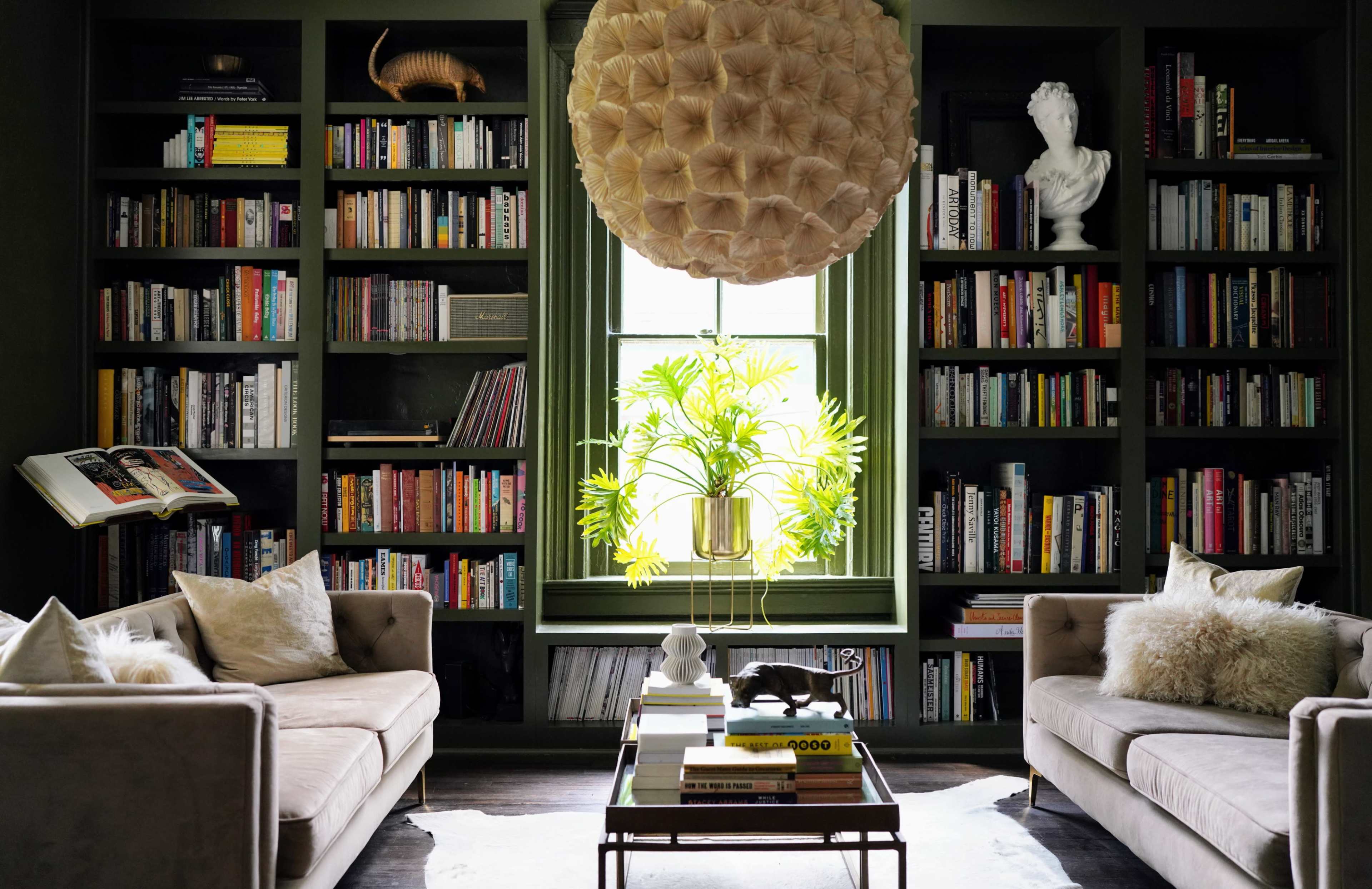 A cozy reading nook features two beige sofas facing a bookshelf filled with books, illuminated by natural light from a large window with a potted plant.