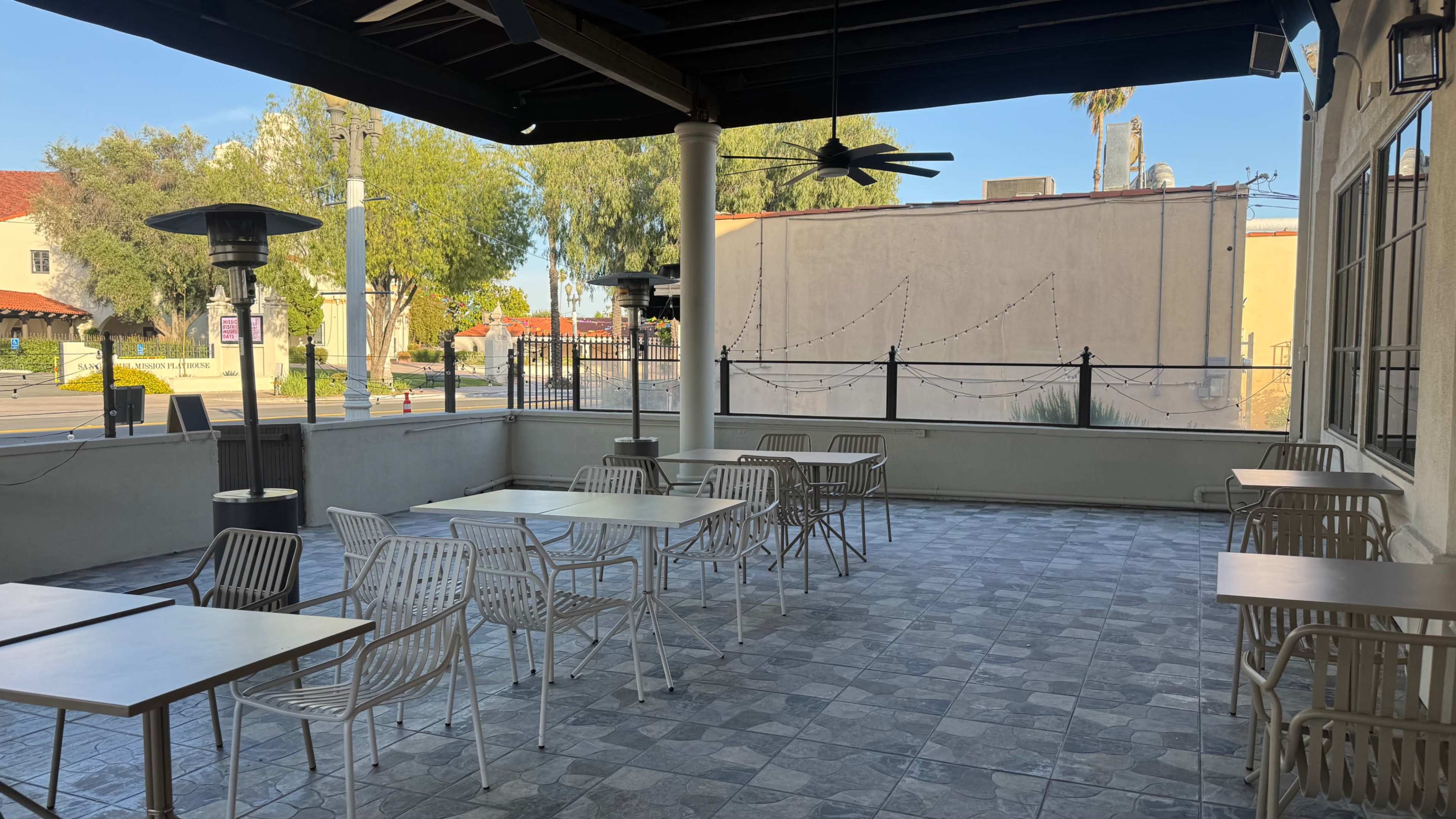 The image shows a partially enclosed outdoor patio area with several white tables and chairs, a ceiling fan, and a view of a street lined with trees and buildings.