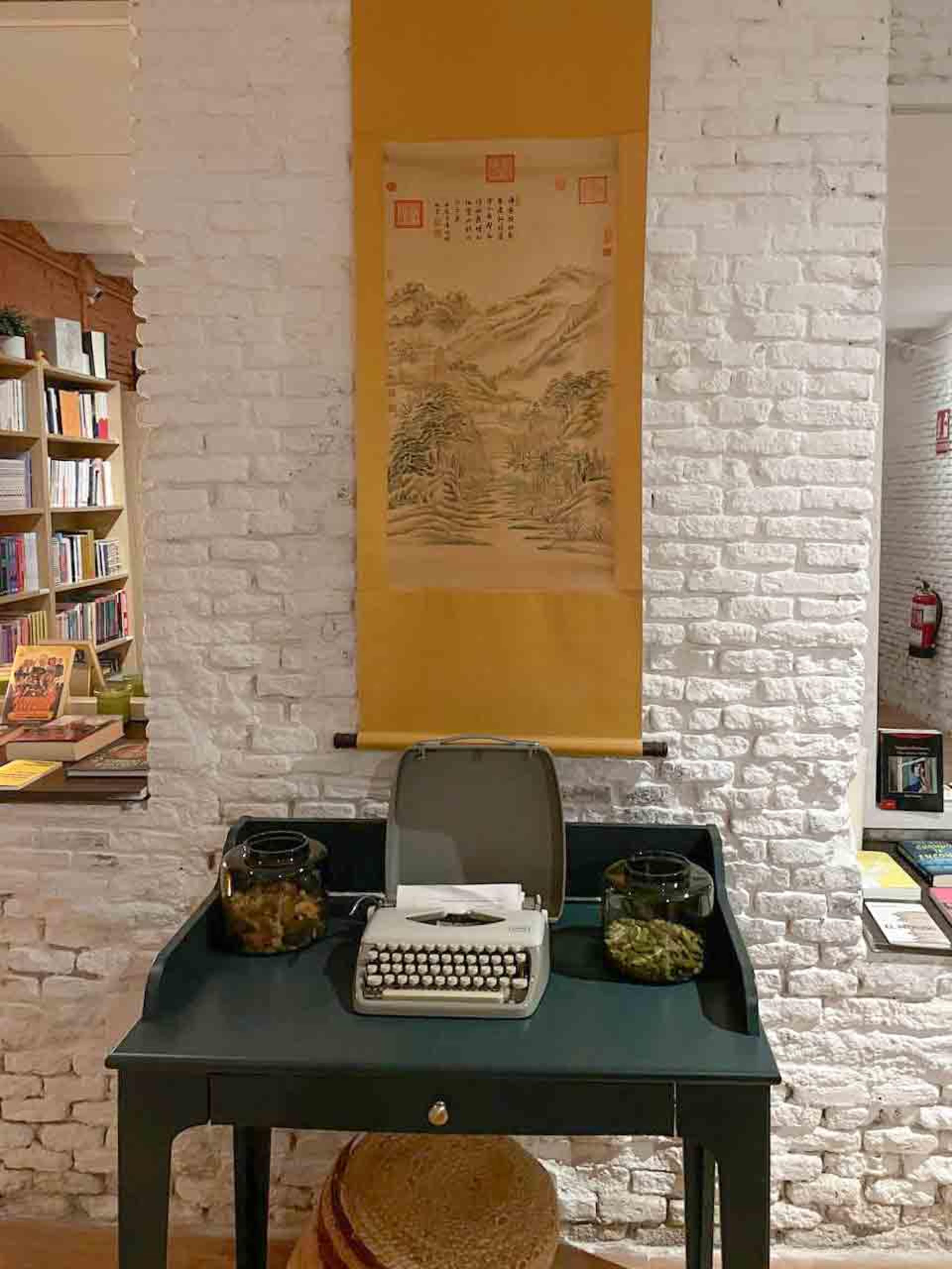 A vintage typewriter is placed on a dark green desk surrounded by jars of preserved items and a hanging scroll featuring a mountainous landscape on a white brick wall.