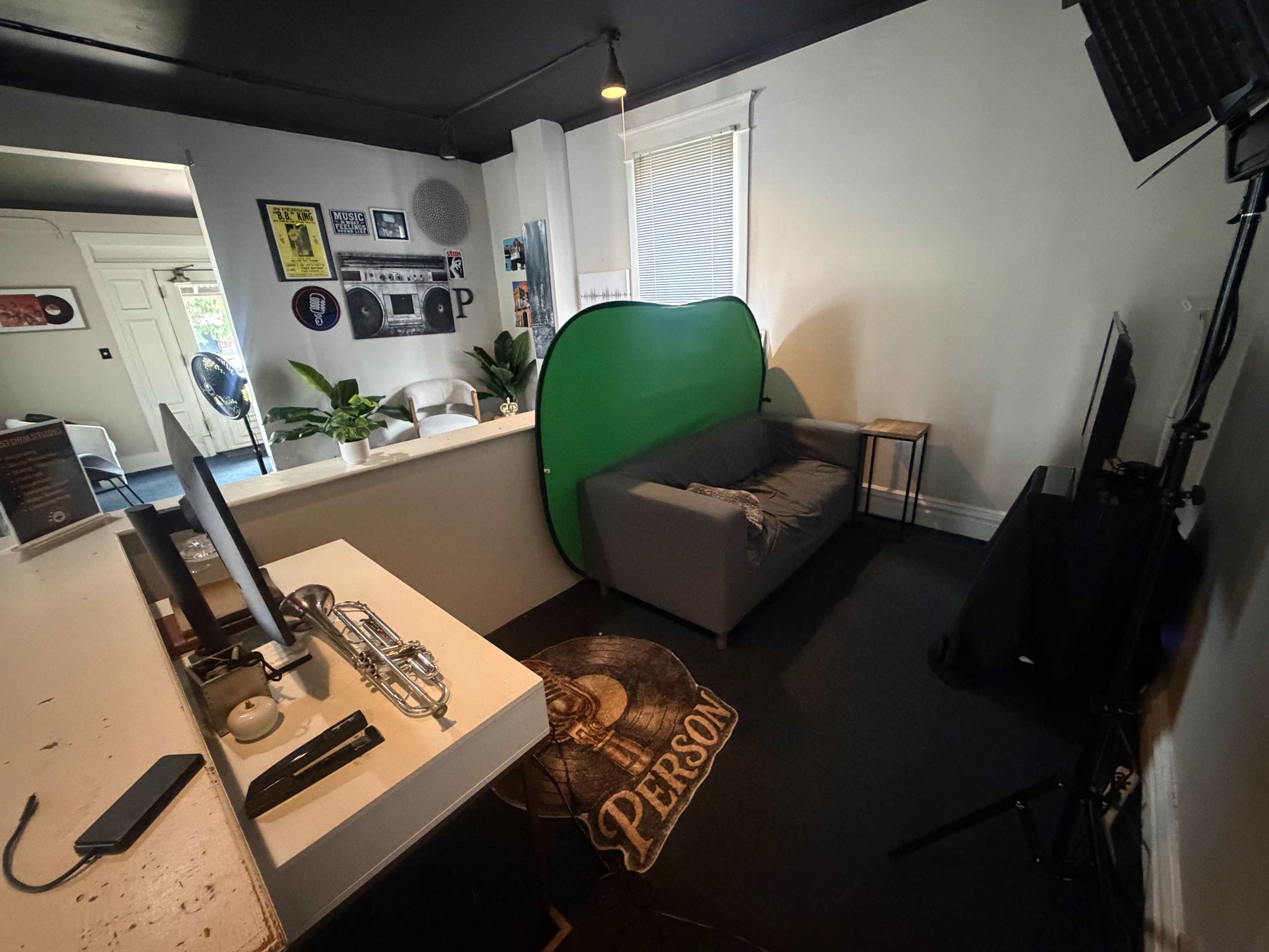 The image shows a small, minimalistic room featuring a green backdrop, a gray couch, a table with a trumpet, and a television setup.